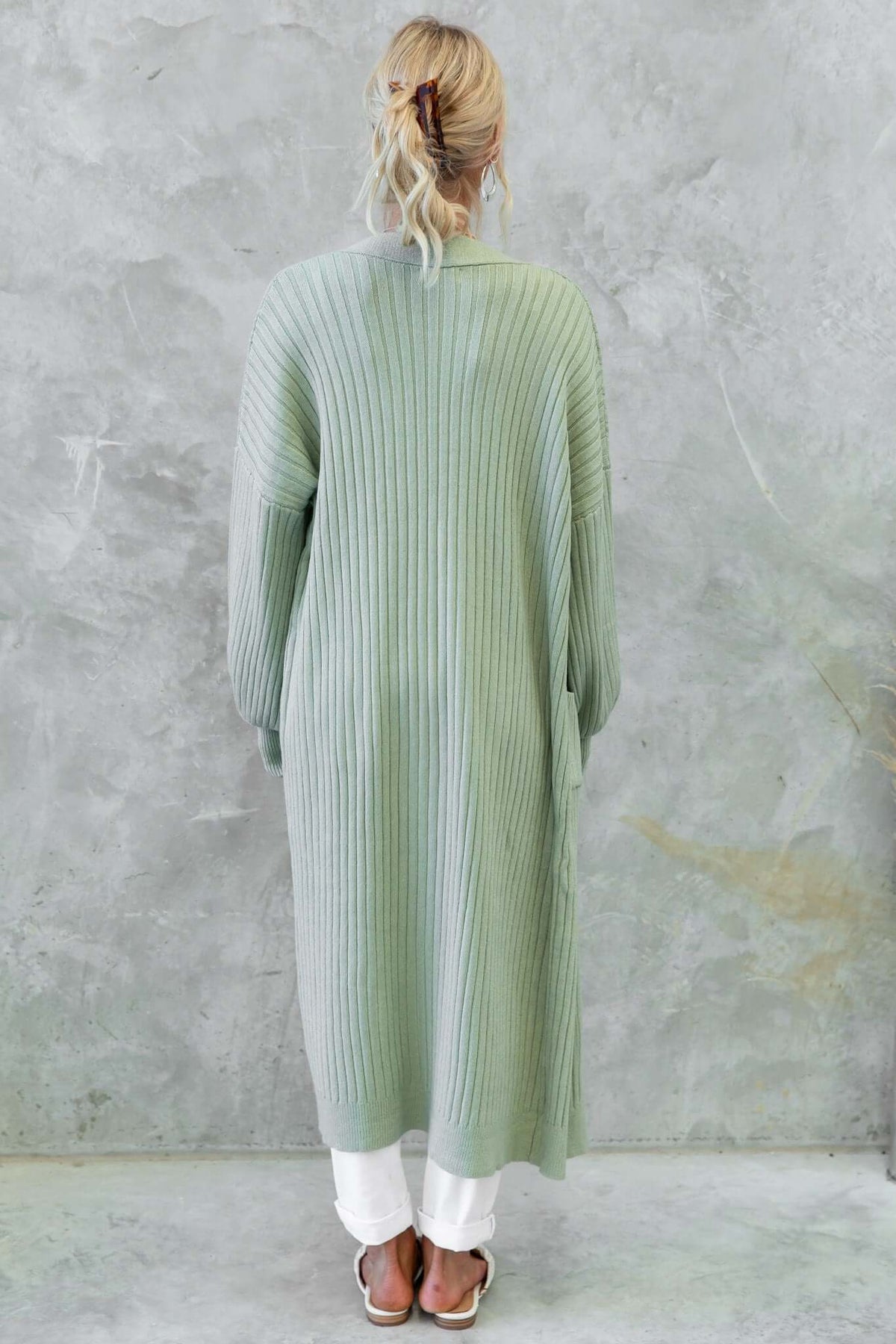 Back view of a longline Fano cardigan in mint green, featuring a ribbed knit design and pockets, styled with white pants.