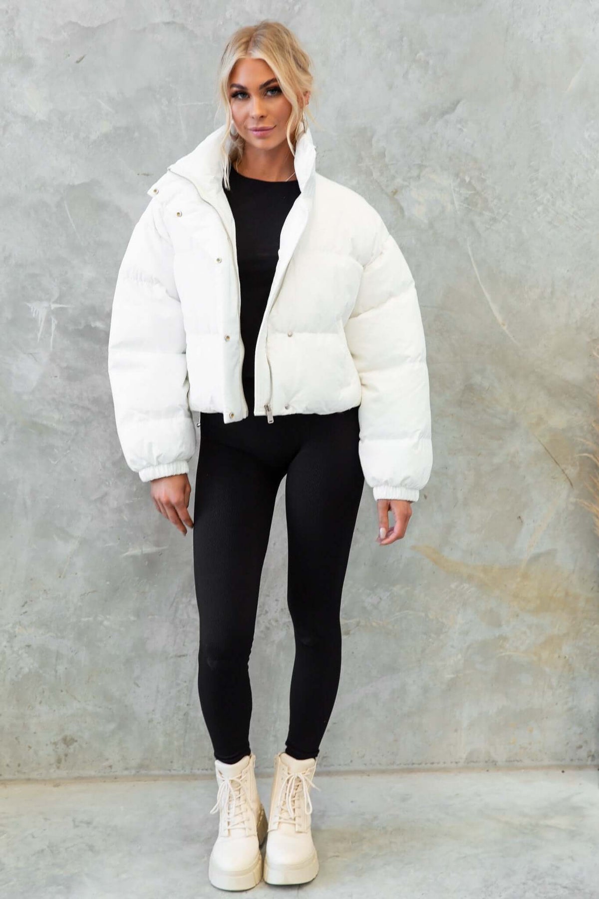 Model wearing white Avril Puffer Jacket with high neckline and adjustable drawstring, styled with black leggings and boots.