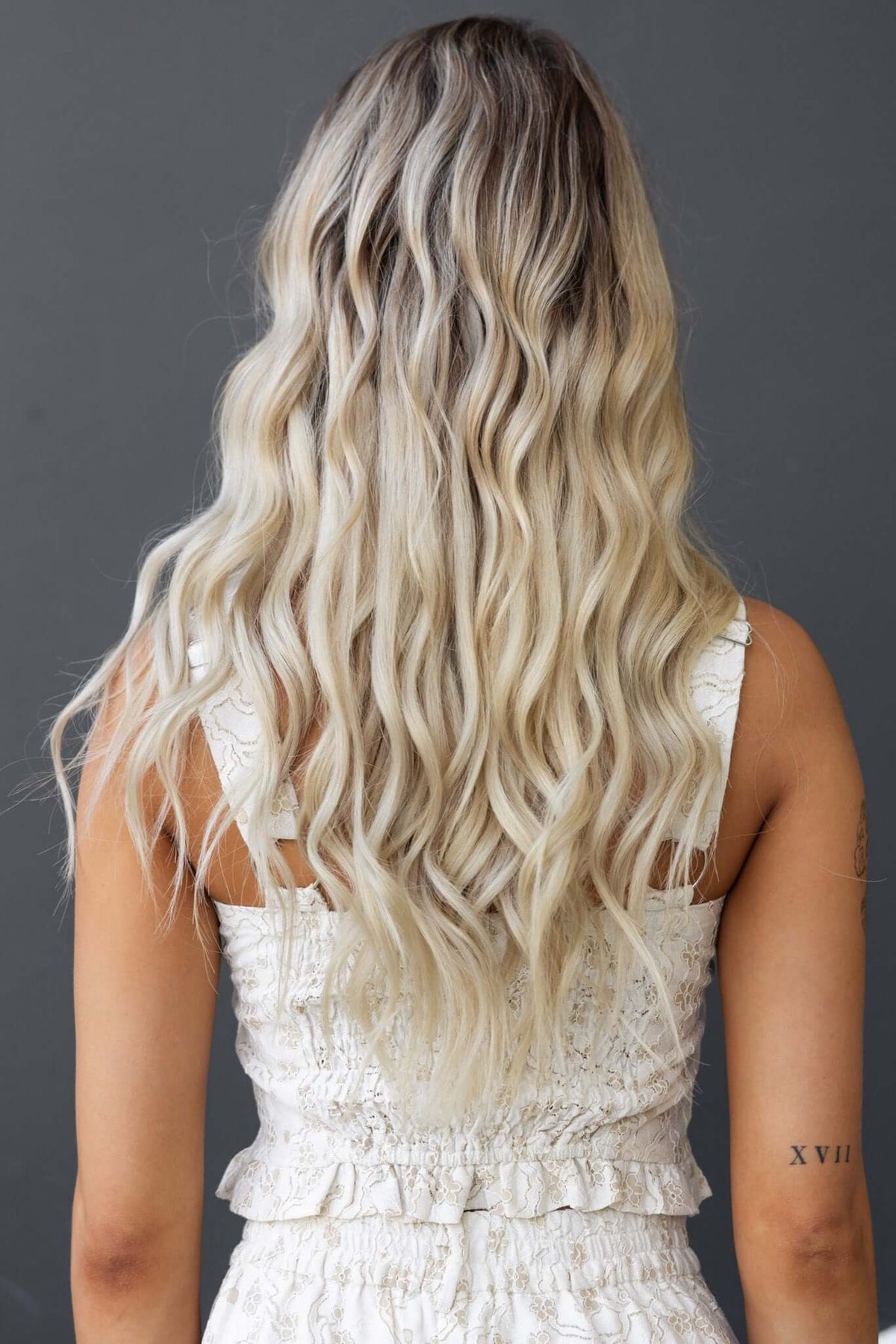 Back view of a woman with long, wavy blonde hair wearing a white textured top.