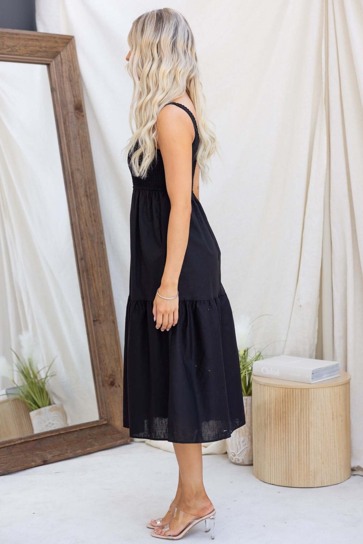 Side view of a woman wearing a black Haidy Midi Dress, featuring an elastic bodice and tiered skirt, styled with a mirror backdrop.
