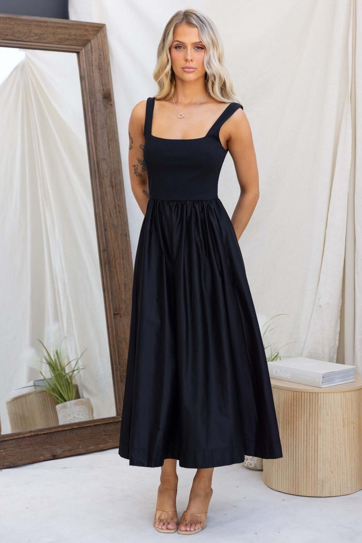 Flint Maxi Dress in black featuring a square neckline, sleeveless design, and full skirt. Model stands with a mirror backdrop.