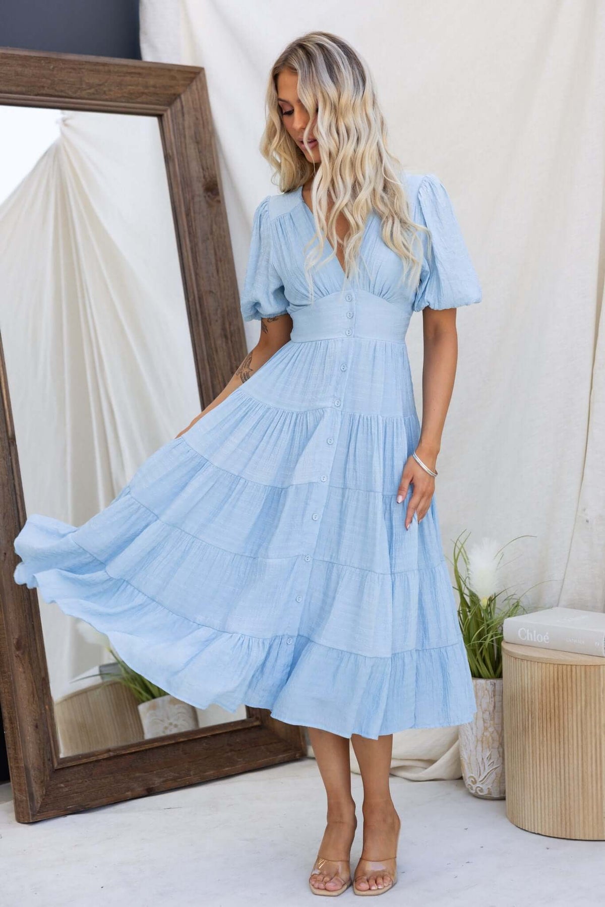 Bindi midi dress in light blue with V-neckline and tiered design, styled with a natural background and reflection.