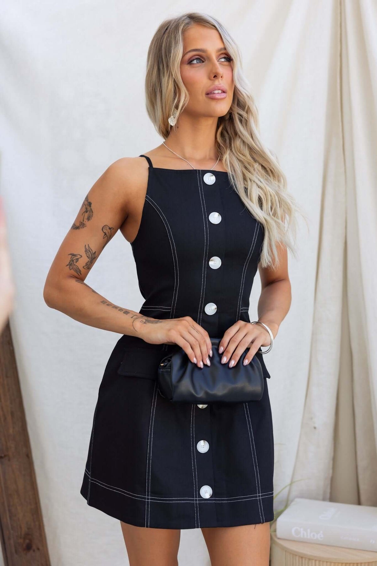Model wearing the Olean Dress, a black sleeveless dress with faux buttons and pockets, stylishly paired with a small handbag.