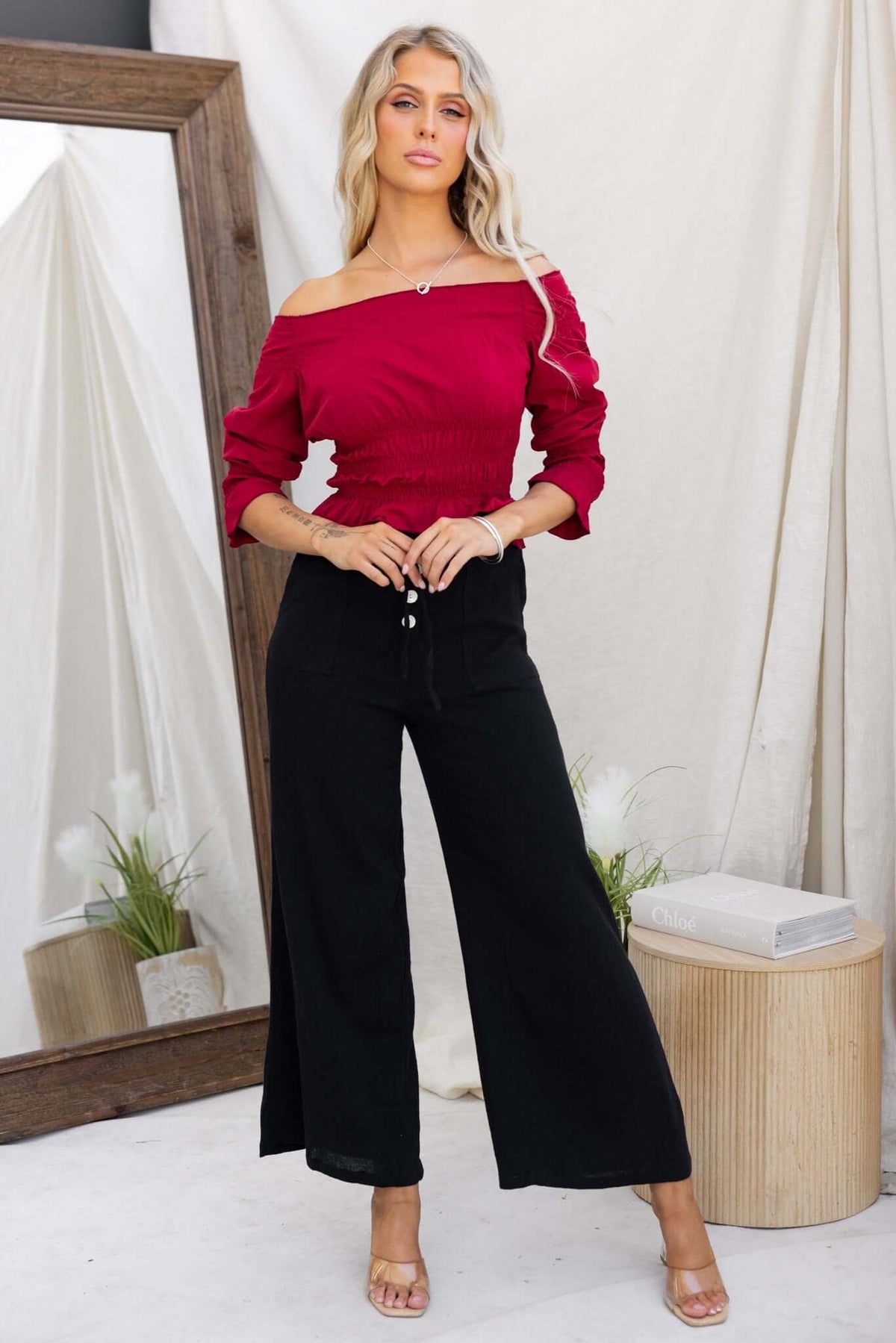 Model wearing a red off-shoulder Jakari Top paired with black wide-leg pants, showcasing a stylish summer outfit.