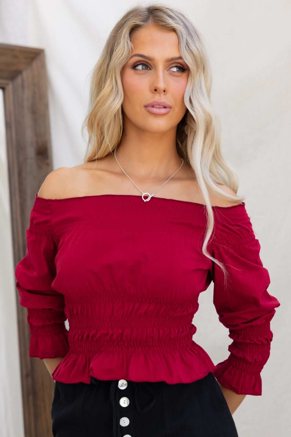 Model wearing a burgundy off-shoulder Jakari Top with elastic features, styled with a necklace and buttoned pants.