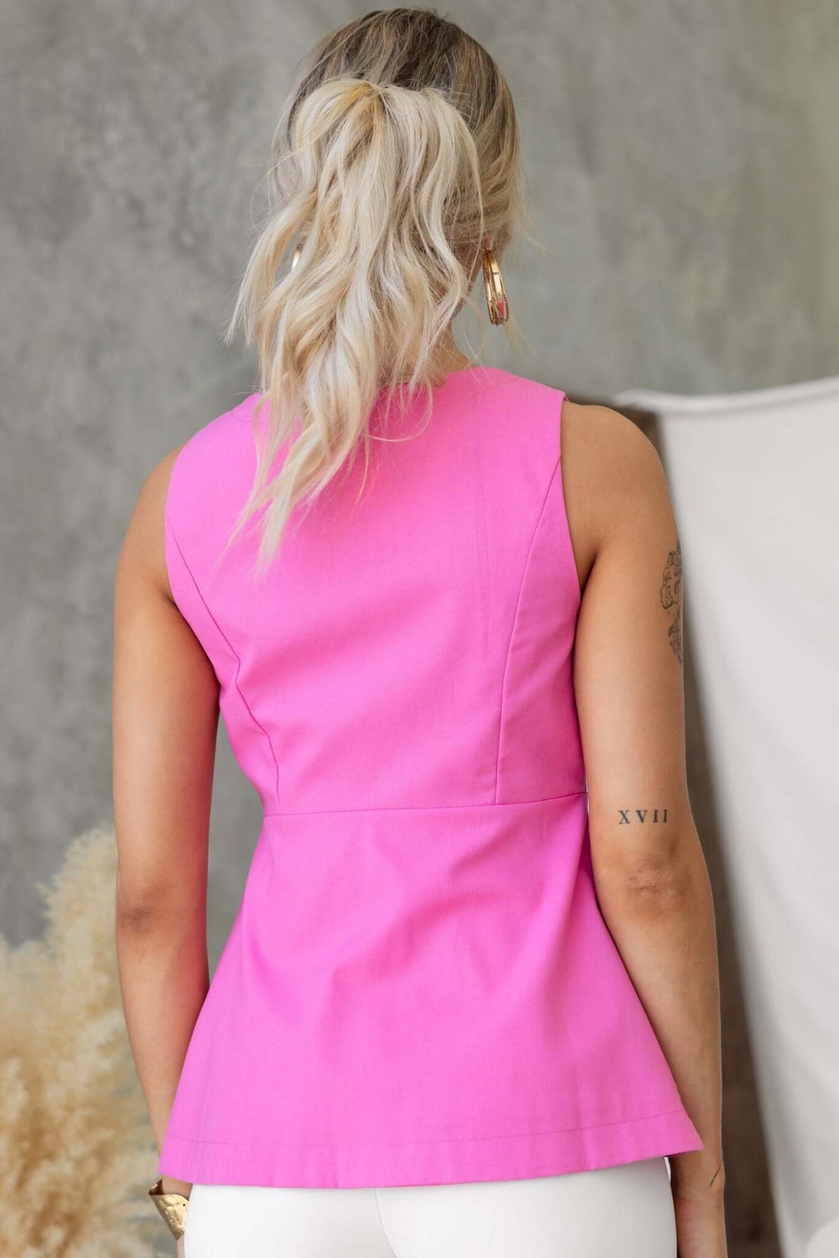 Back view of a woman wearing a pink vest top featuring a peplum design and no sleeves, styled with white pants.