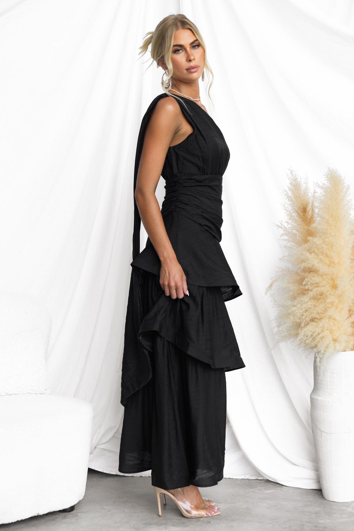 Elegant black one-shoulder maxi dress with ruffle hem and draping detail, styled with a soft background.