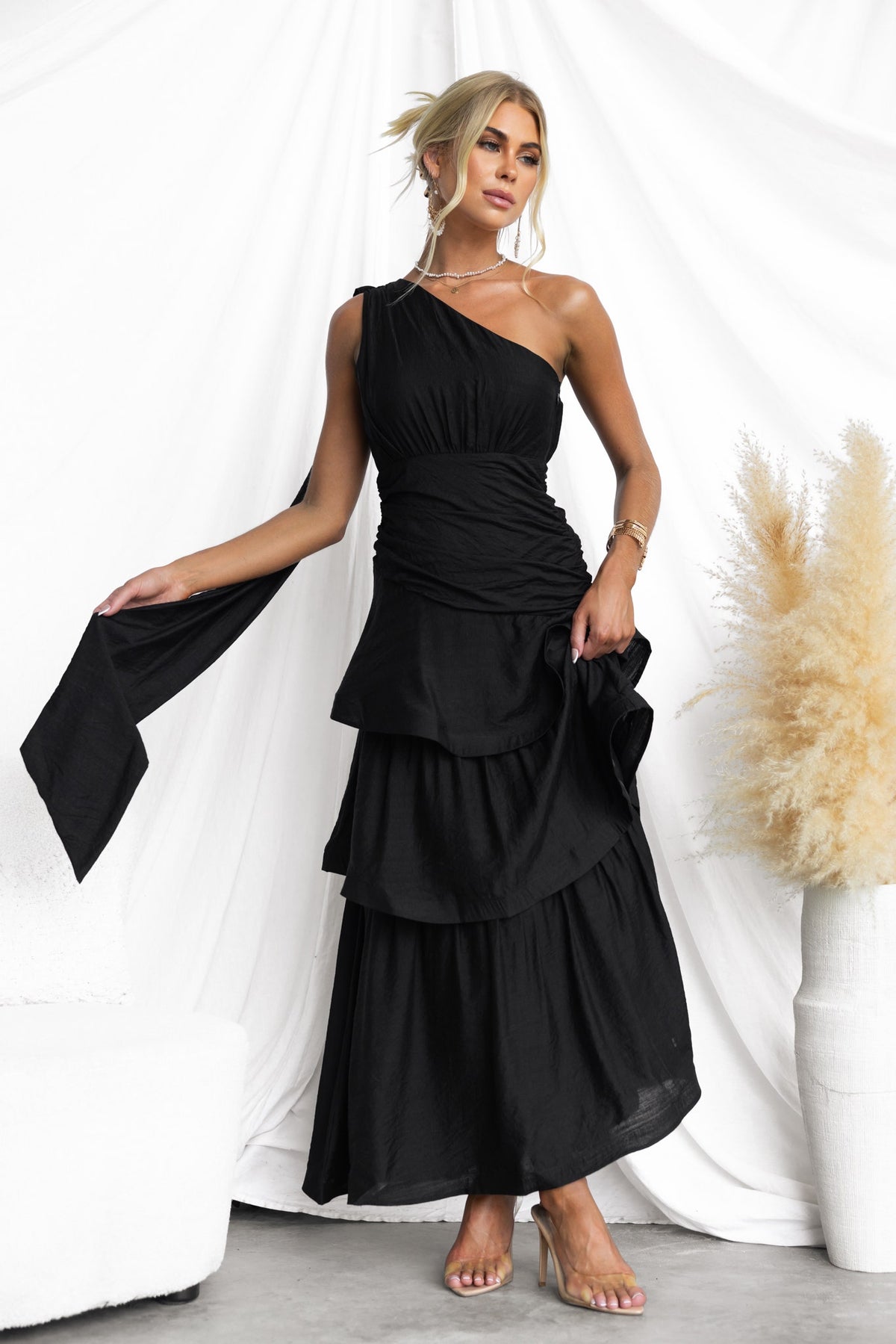 Model wearing a black one-shoulder maxi dress with draped straps and ruffled hem, styled in an elegant setting.