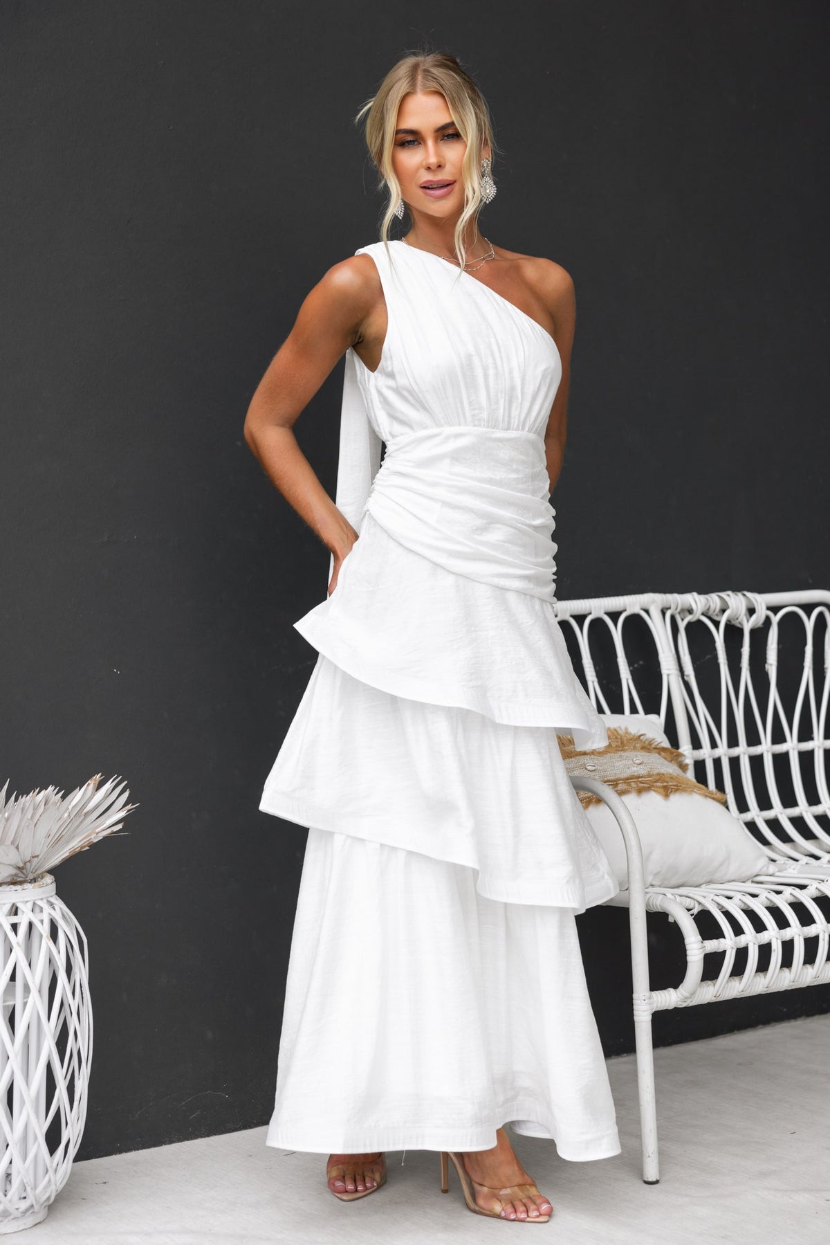 Model wearing a stylish white one-shoulder maxi dress with ruffle hem and tie detail, posed against a dark background.