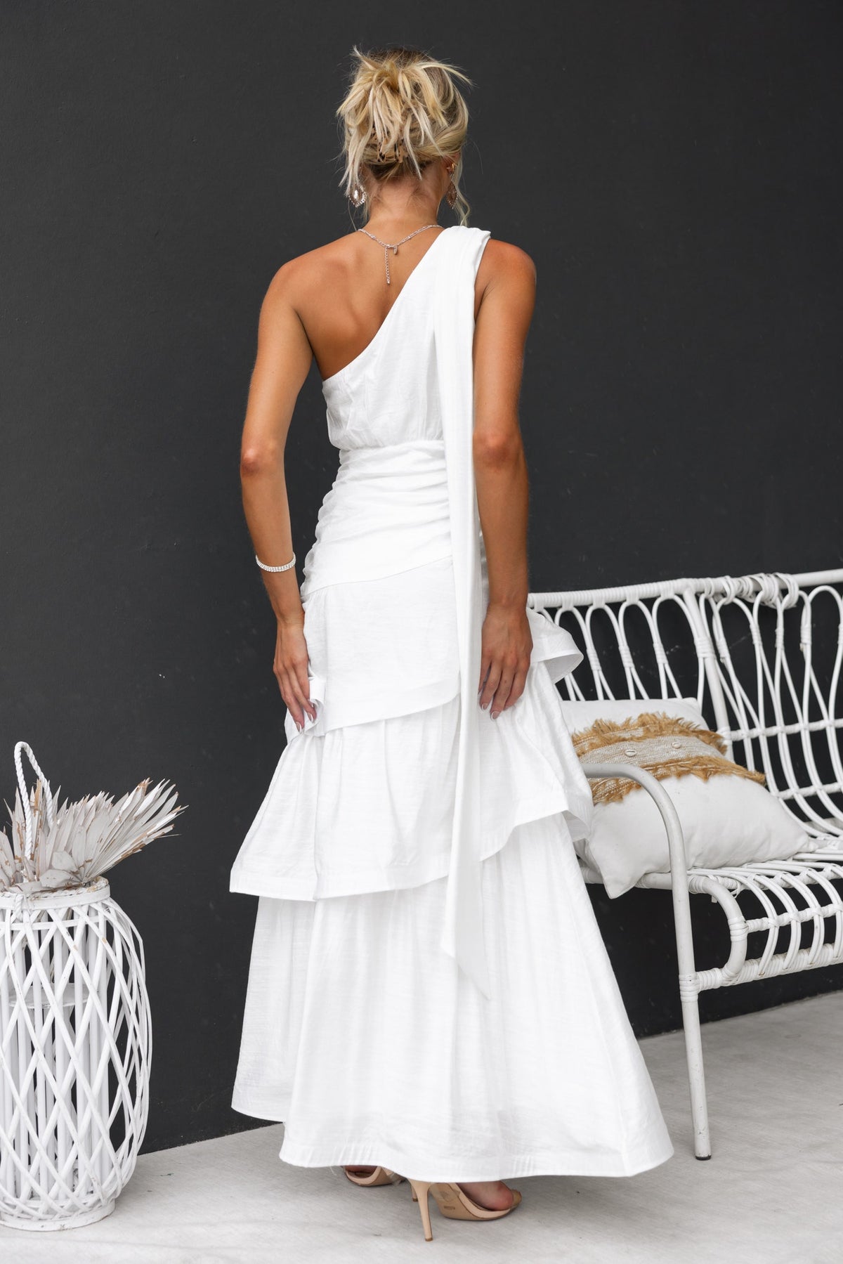 Elegant white maxi dress with one-shoulder neckline and ruffled hem, displayed against a modern backdrop.