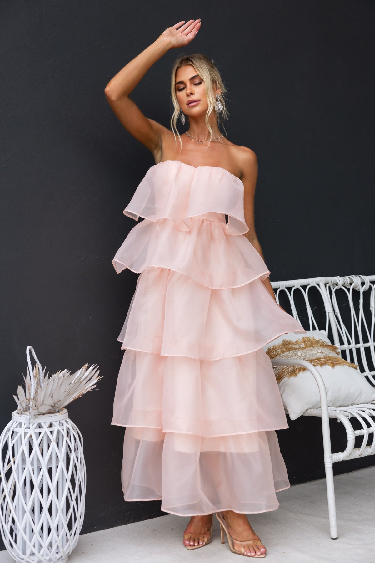 Lilianna maxi dress in soft peach, strapless with tiered chiffon layers, model poses elegantly beside a white chair.