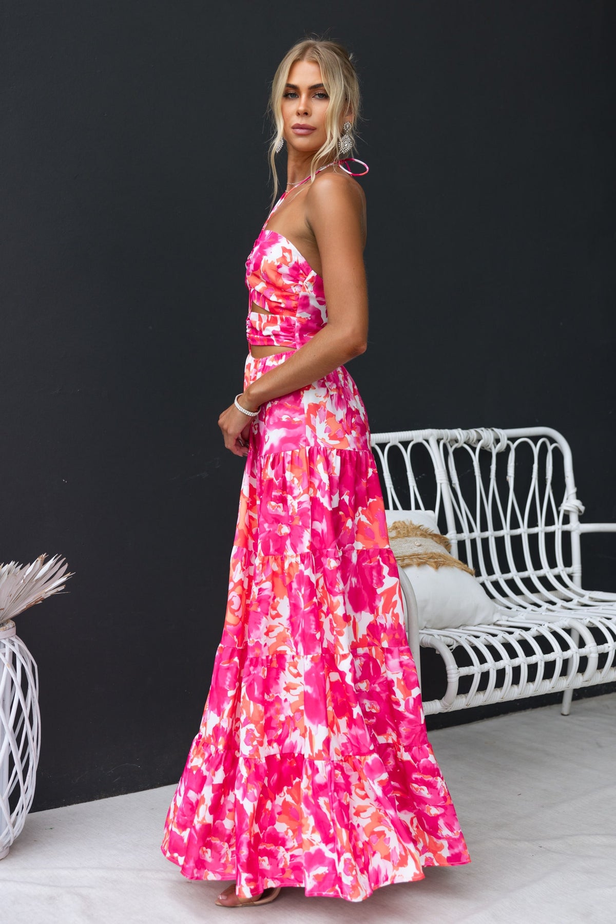 Woman in pink floral print Lauri maxi dress with a tie halter neckline and cut-outs, standing elegantly against a dark background.