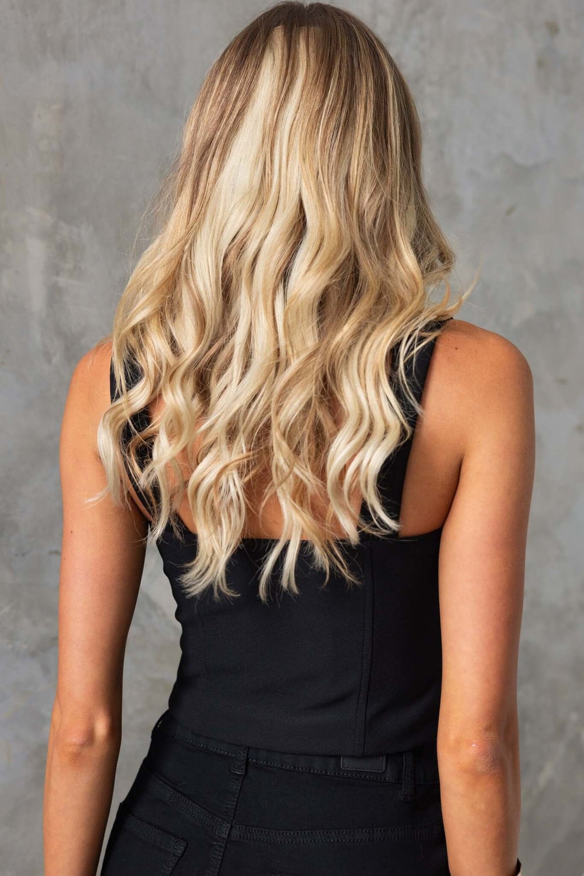 Back view of a model showcasing the Qlair Top in black with wavy blonde hair against a textured background.