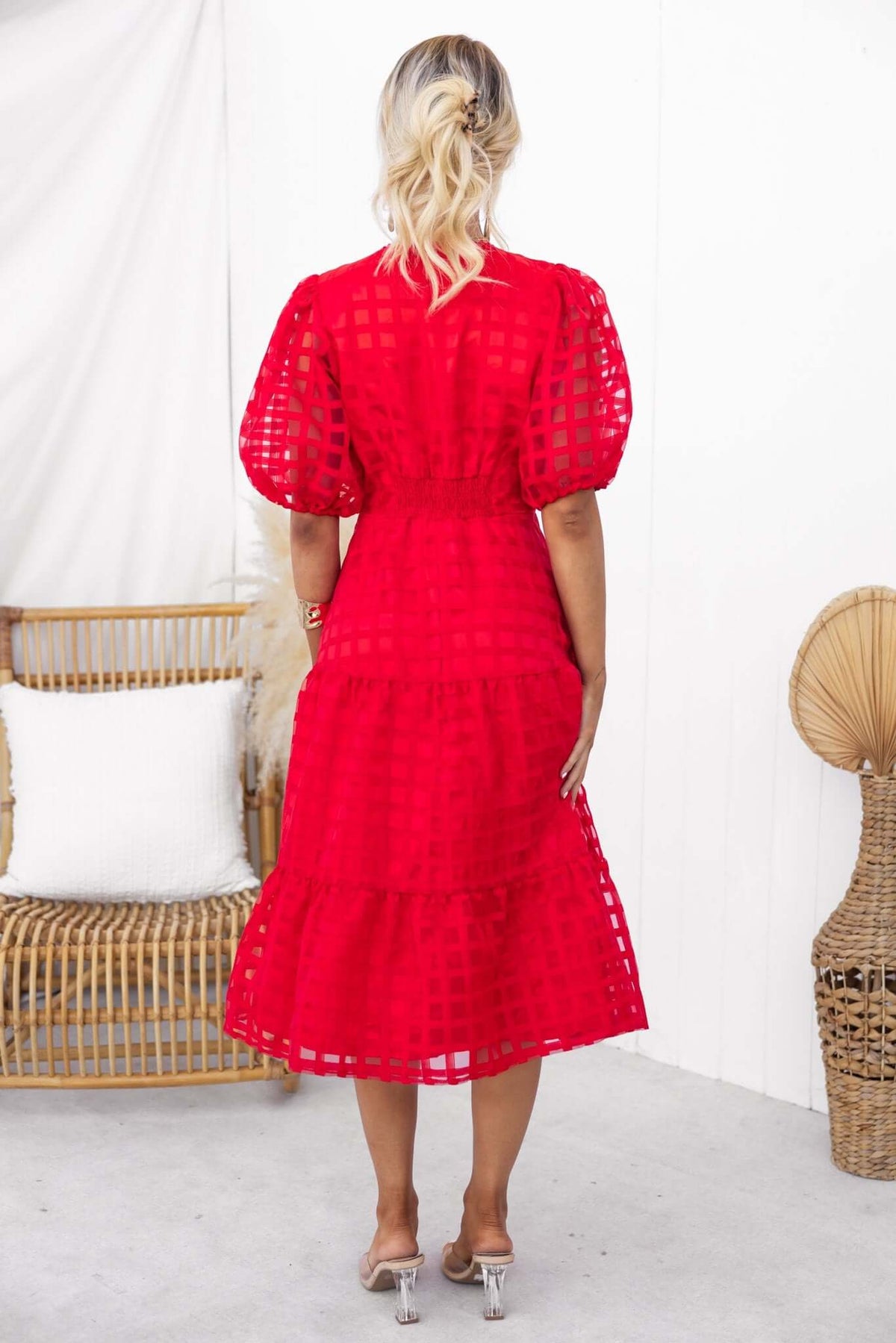 Model wearing red Barker Midi Dress from the back, featuring puff sleeves and tiered design, styled with clear heels.