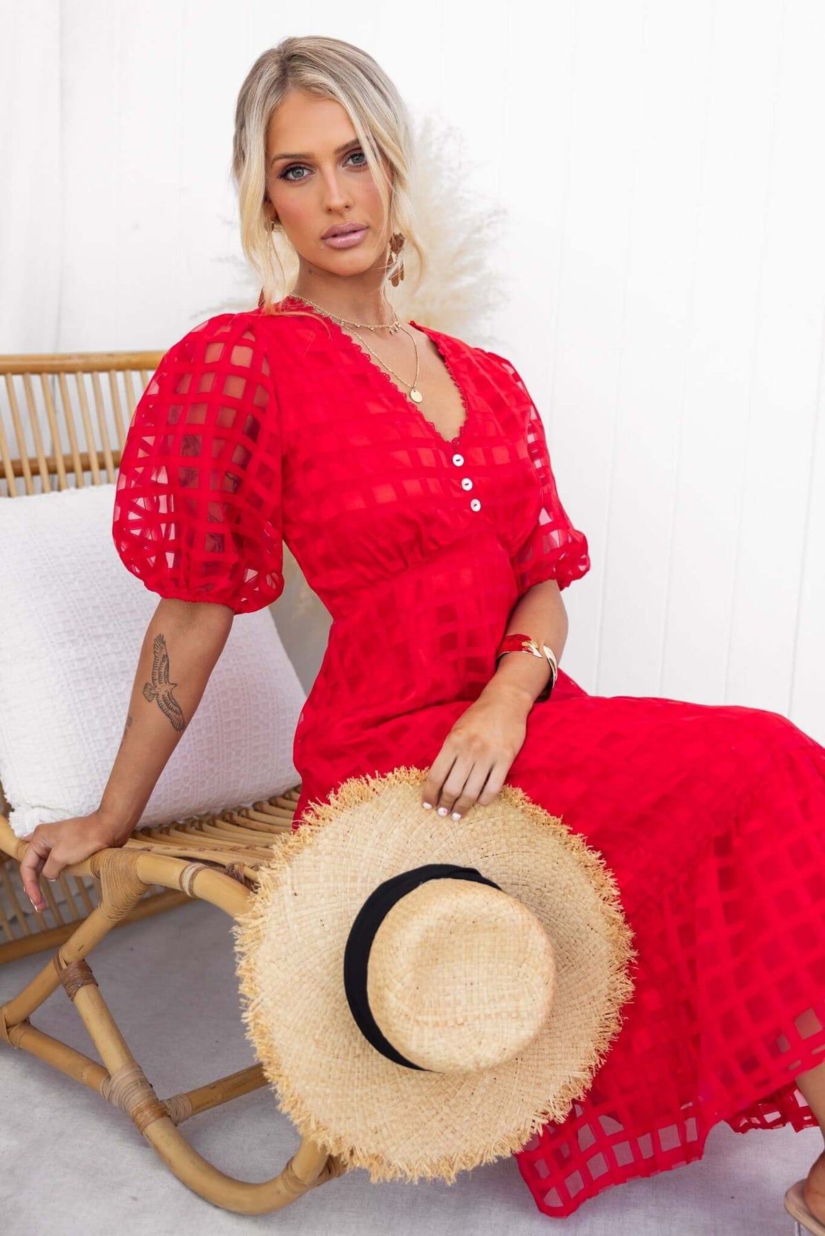 Model wearing a red Barker Midi Dress with puff sleeves, sitting on a chair, holding a straw hat.