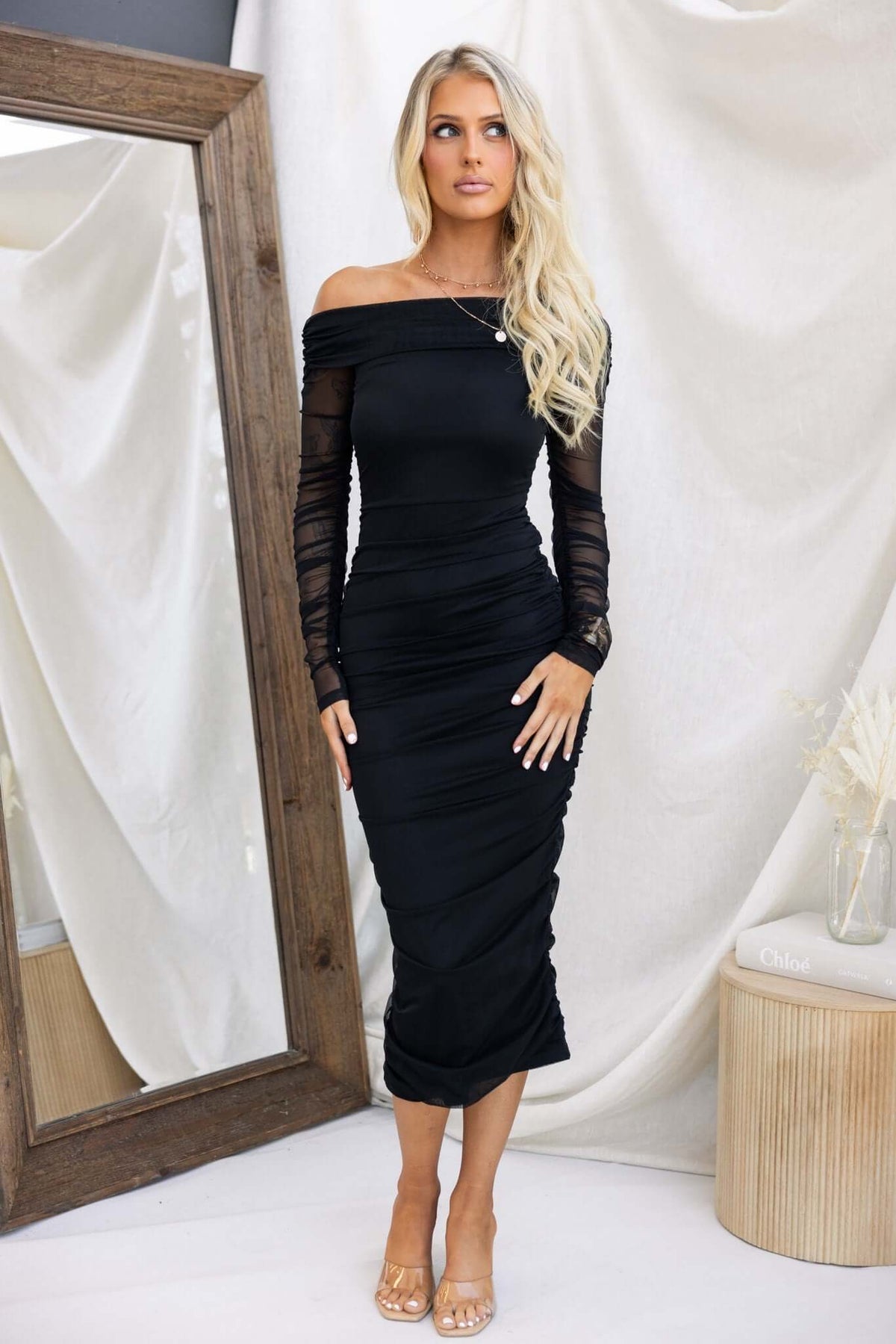 Elegant off-shoulder Hilton Midi Dress in black, featuring a fitted design and long sheer sleeves, styled with open-toe heels.