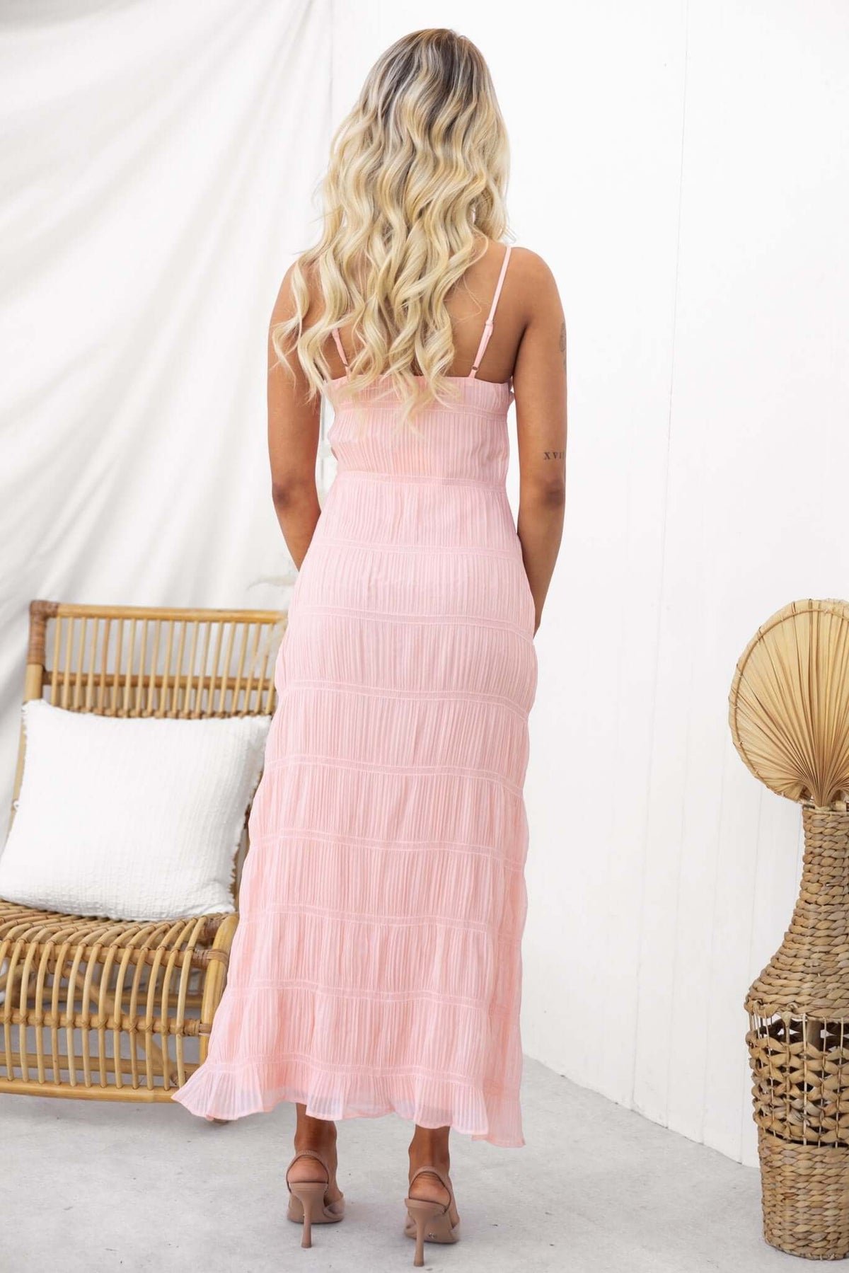 Back view of a woman in a pink maxi dress with thin straps, styled with soft waves and a stylish decor backdrop.