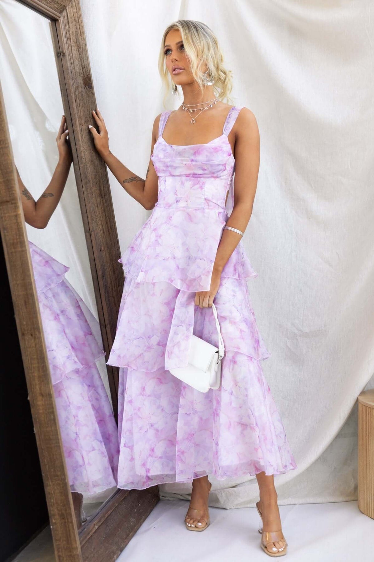 Model wearing a Bennett Ave Maxi Dress with tiered frills and a lilac floral design, standing next to a mirror.