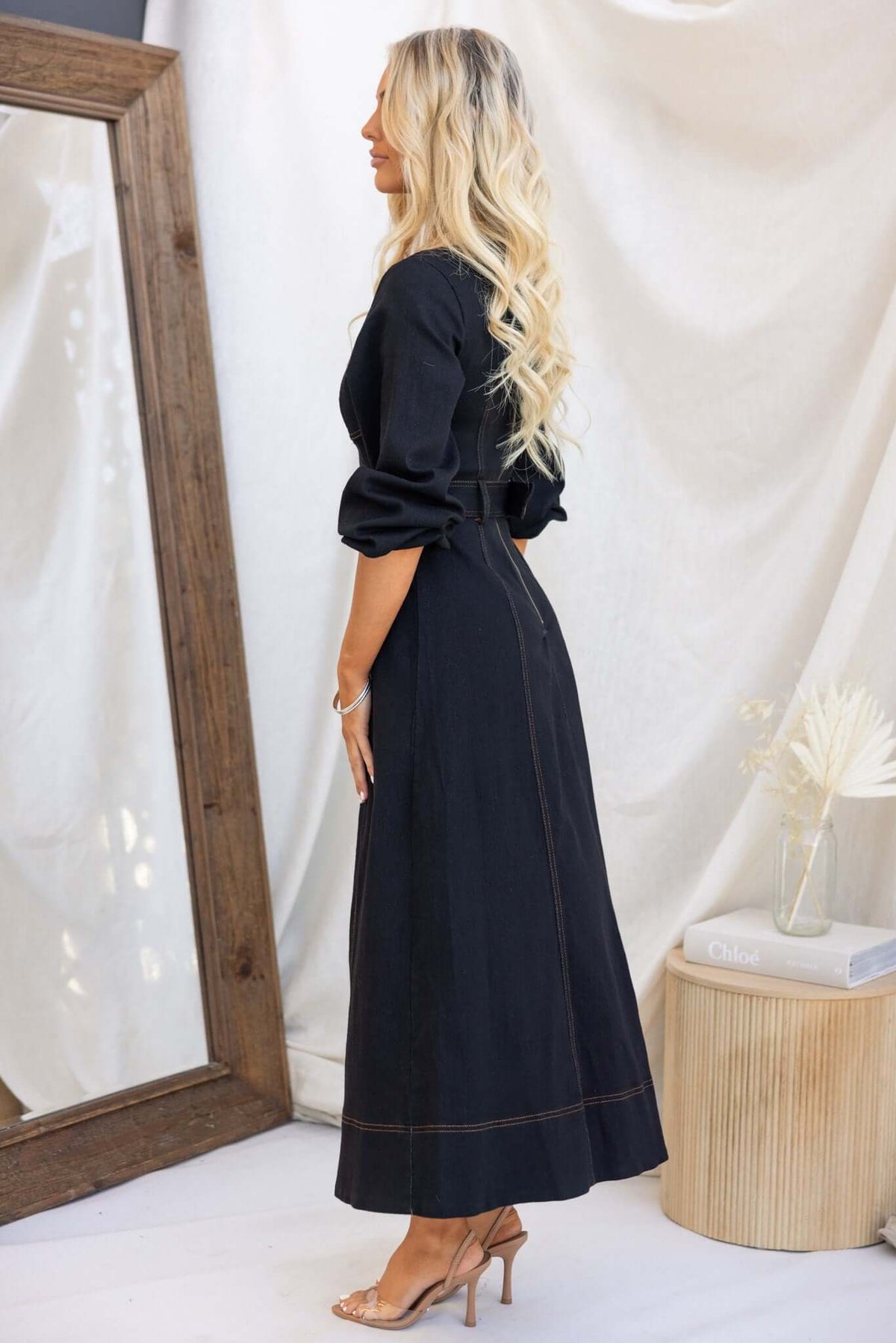 Side view of a woman in a black denim maxi dress featuring faux buttons and puff sleeves.