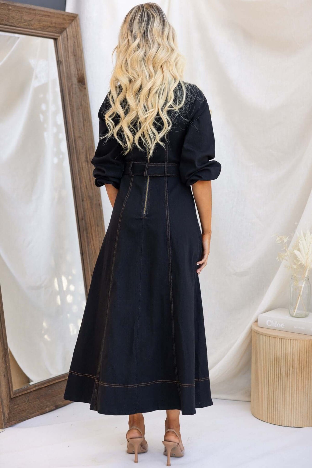 Back view of a woman in a black denim maxi dress with faux buttons, elastic sleeves, and a removable belt.