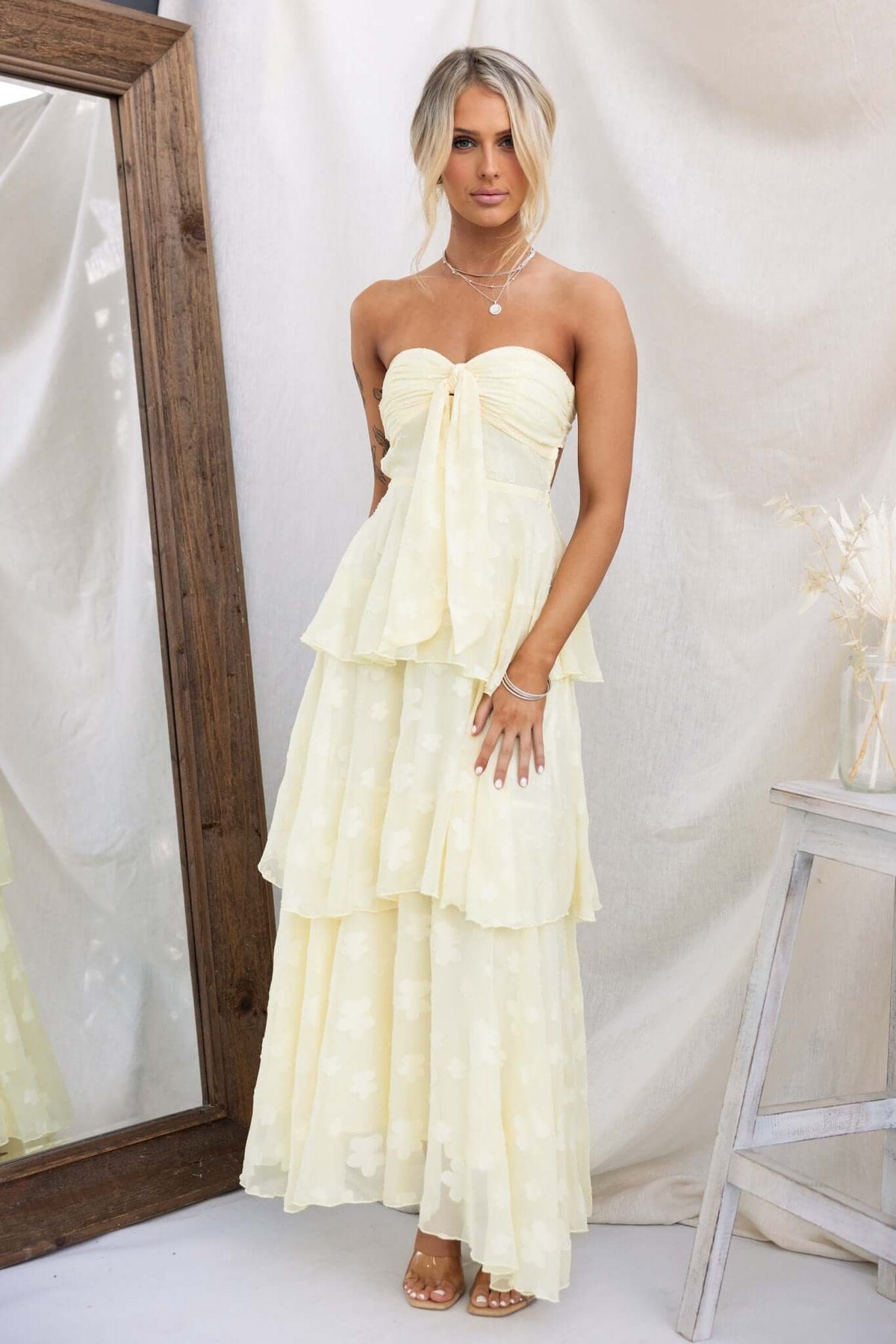Model wearing a soft yellow Soft Whispers Maxi Dress with a tiered ruffle skirt in front of a mirror.