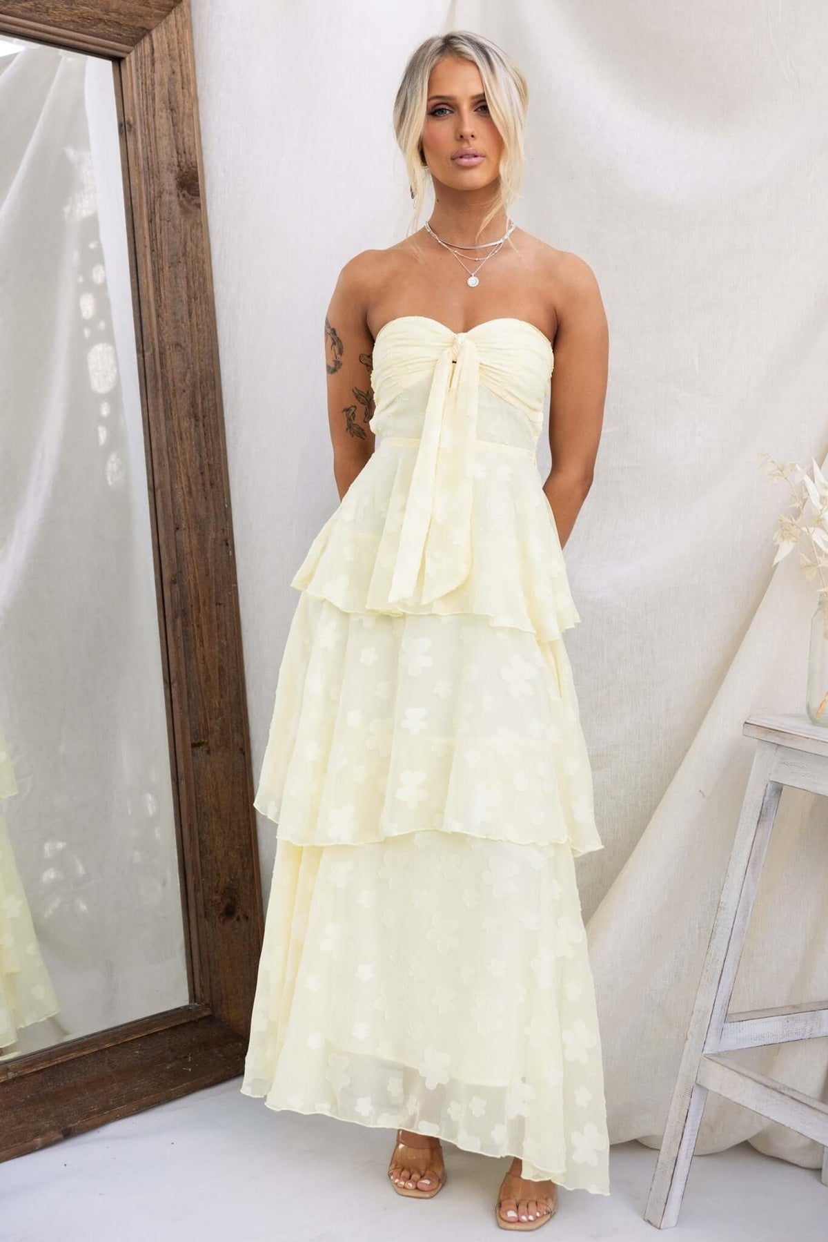 Model wearing a soft yellow tiered ruffle maxi dress with a tie at the bust, showcased against a rustic mirror.