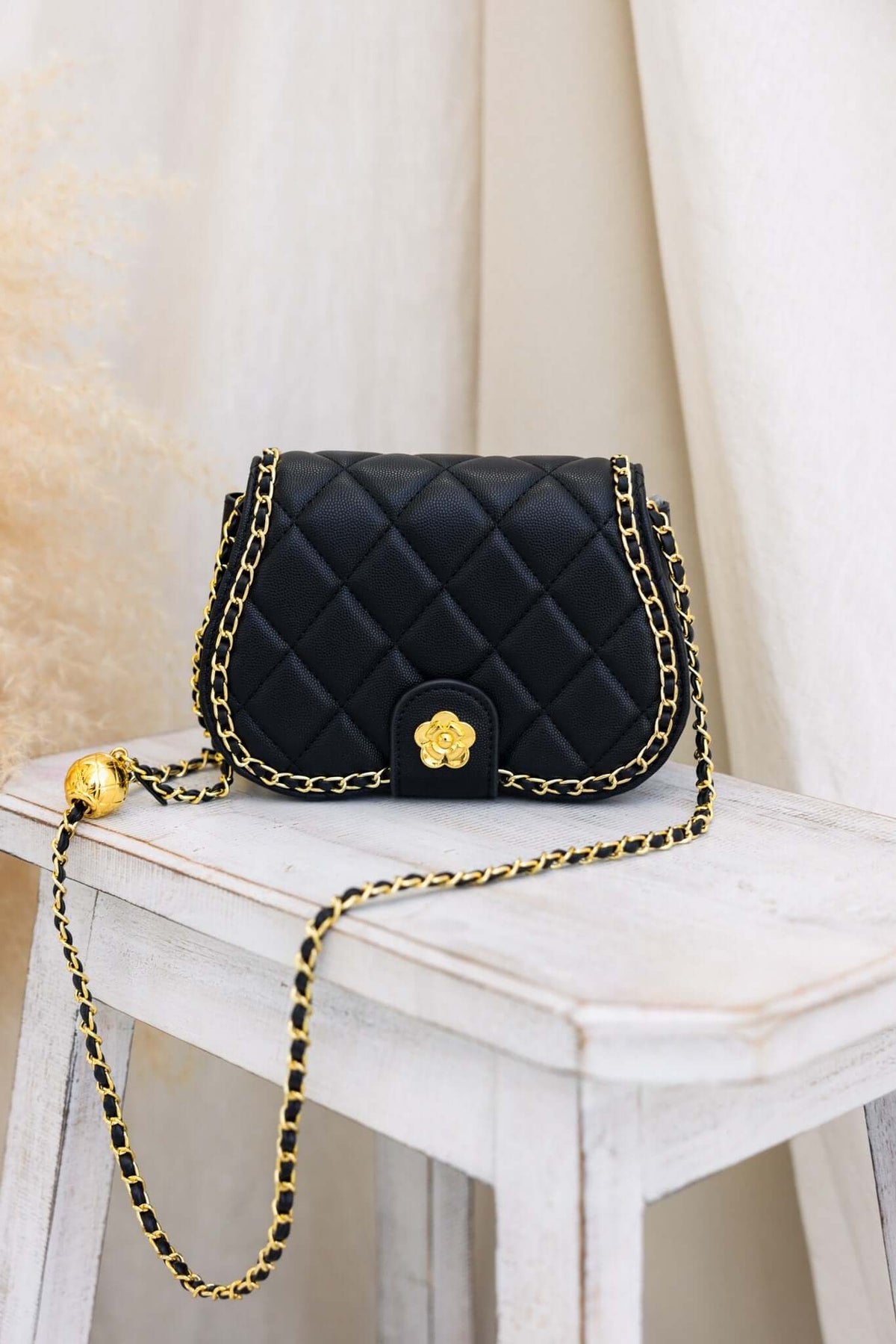 Chic black quilted handbag with gold chain and floral embellishment, placed on a white wooden table.