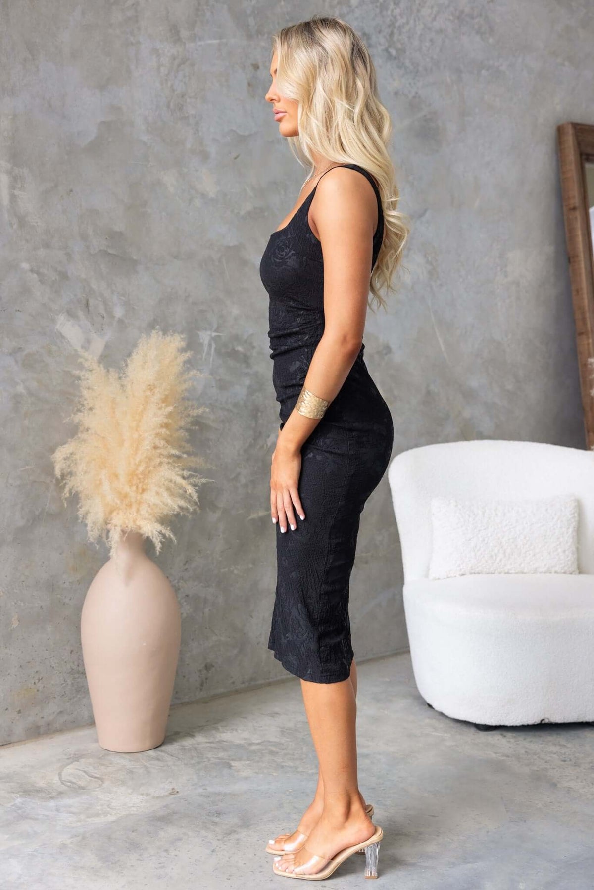 Side view of a woman in a black Sheridan Midi Dress with ruched sides, showcasing textured material and a fitted silhouette.