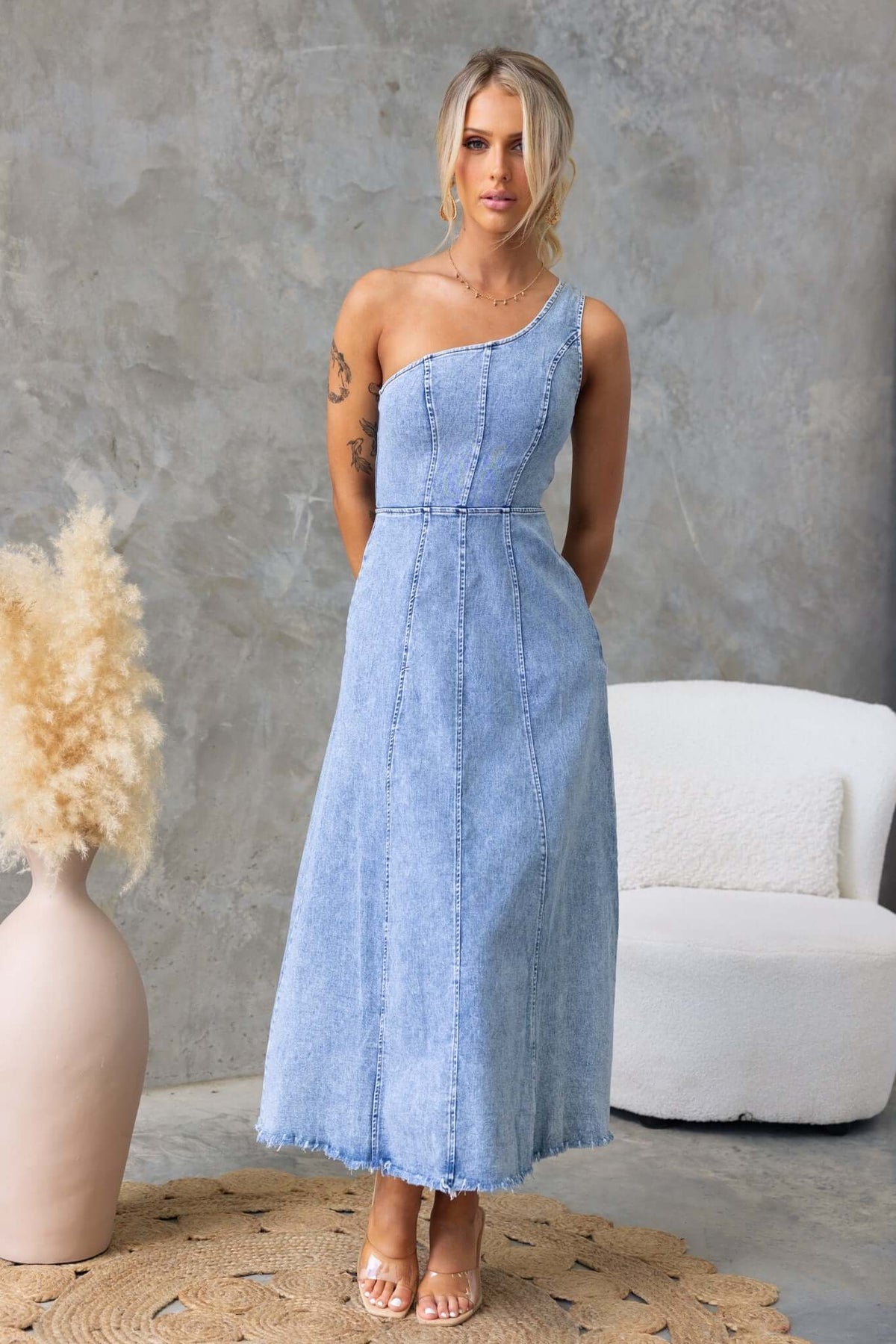 One-shoulder denim maxi dress with removable brooch and raw hemline, styled elegantly in a modern setting.