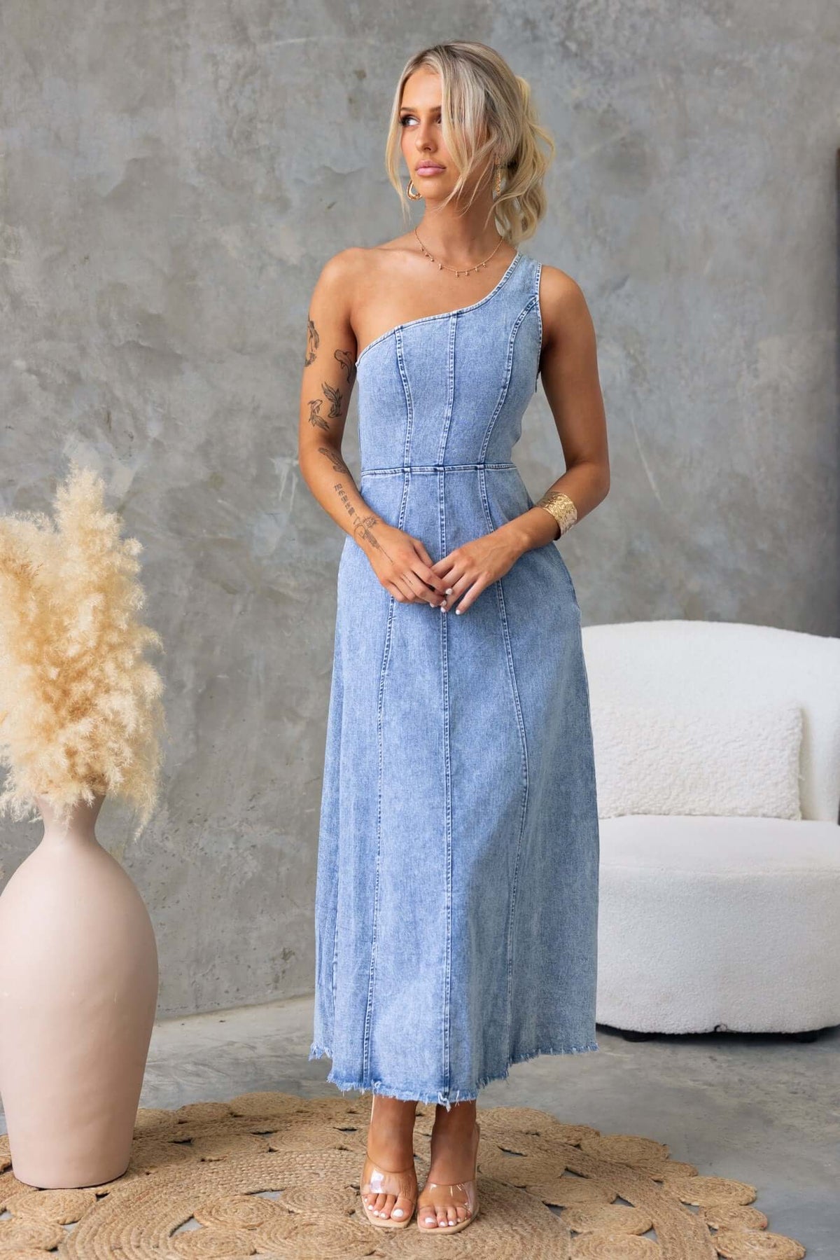 One-shoulder denim maxi dress with removable brooch, featuring a raw hemline and stretchy material, styled with a neutral background.