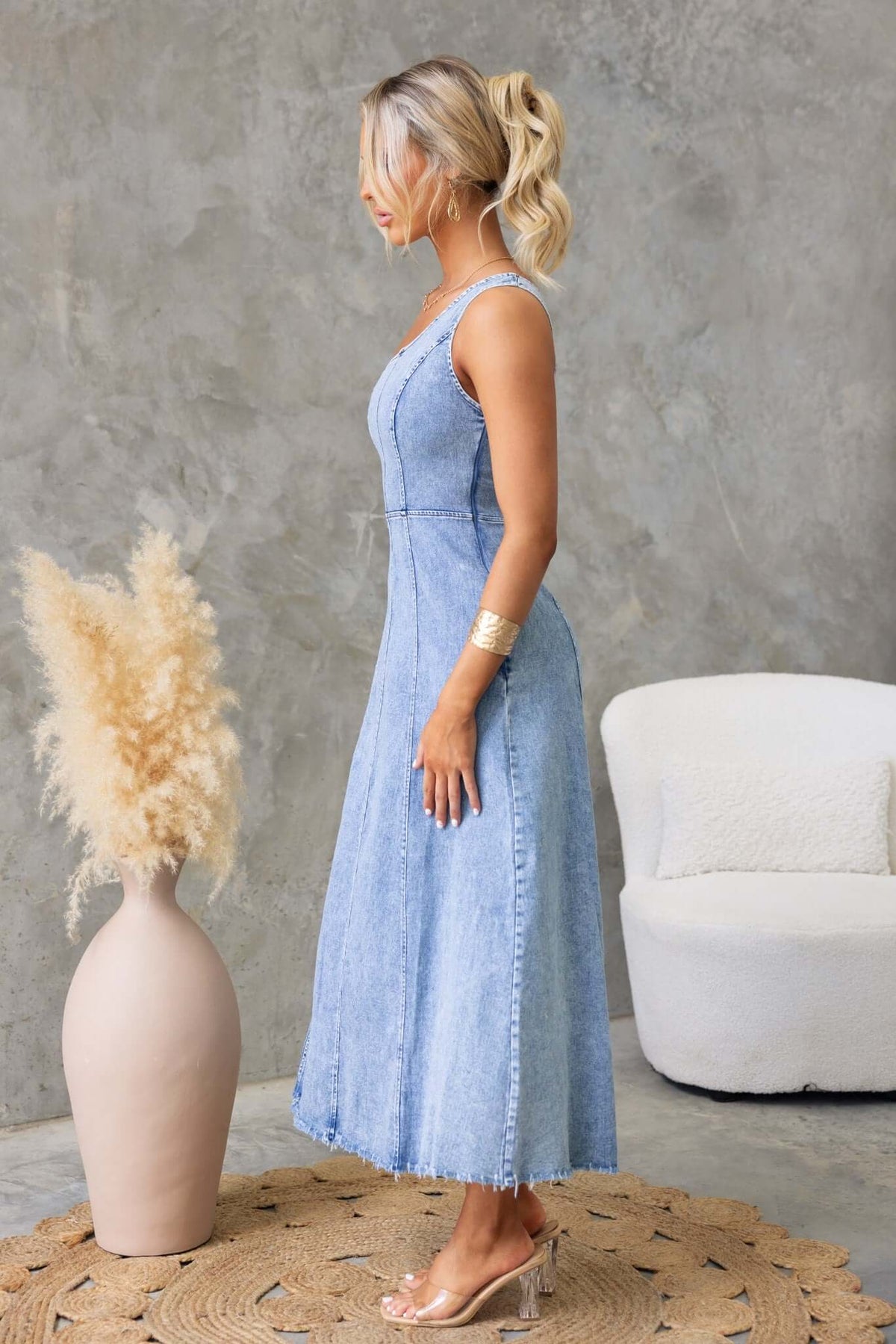 Side view of a woman in a denim maxi dress with one-shoulder neckline and raw hemline, sitting in a modern interior.