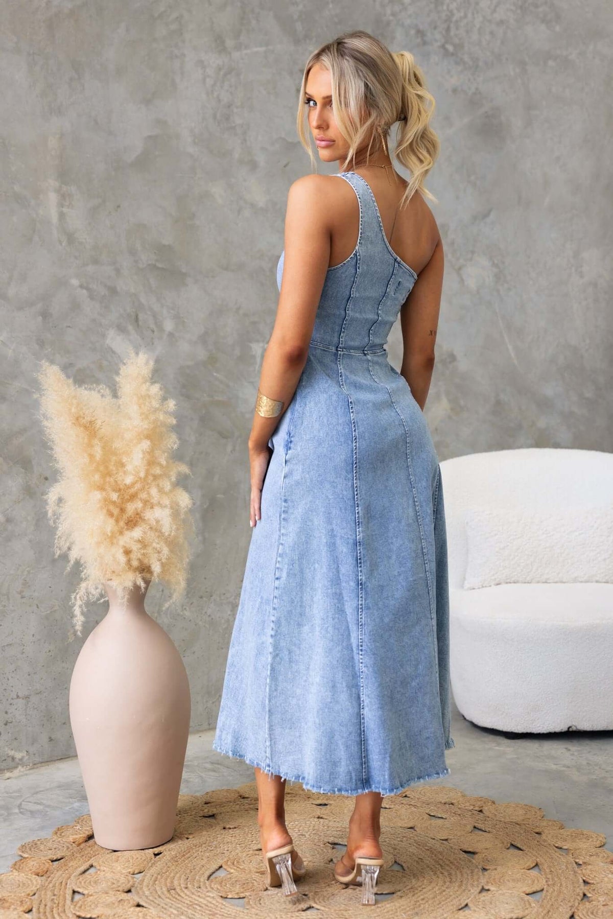Back view of a woman in a Duet Denim Maxi Dress with raw hemline and one-shoulder neckline, styled with a minimalist background.