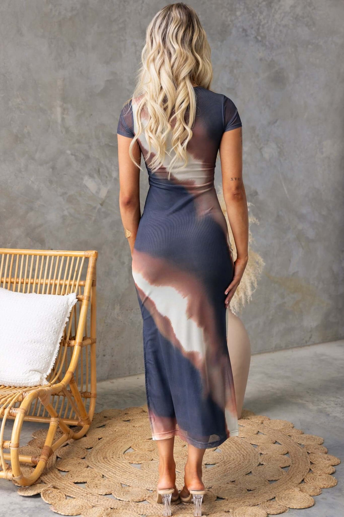 Back view of Dusk Maxi Dress with mesh overlay and side split, styled with wavy hair, against a textured background.