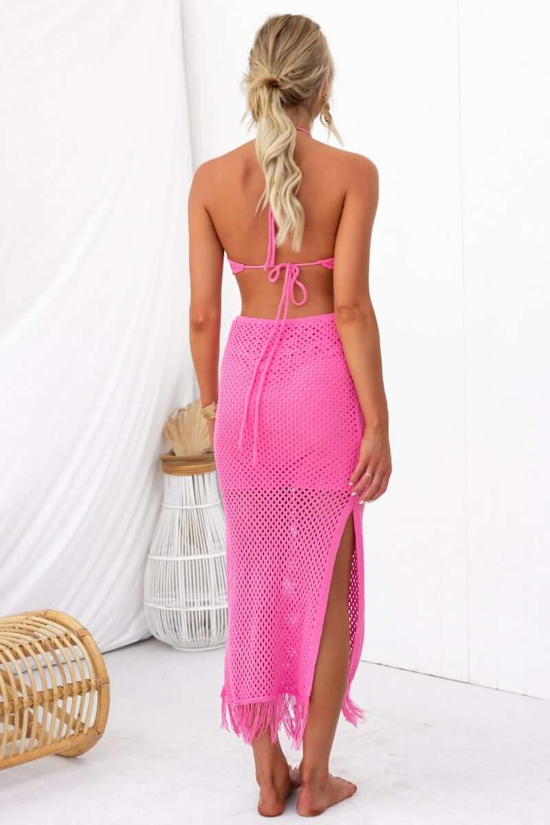 Back view of a model in a bright pink crochet dress with cutouts and a high slit, paired with a matching halter top.