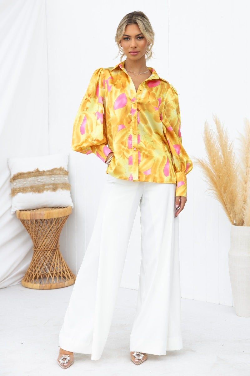 Model wearing Indianna Top, a satin button-up shirt with colorful print, paired with white wide-leg pants.