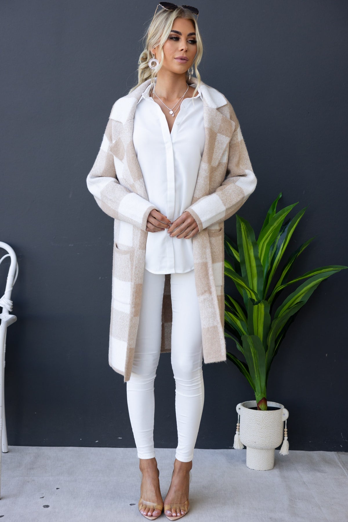 Model wearing a longline Fatale Cardigan with pockets over a white shirt and white pants, styled with a plant backdrop.