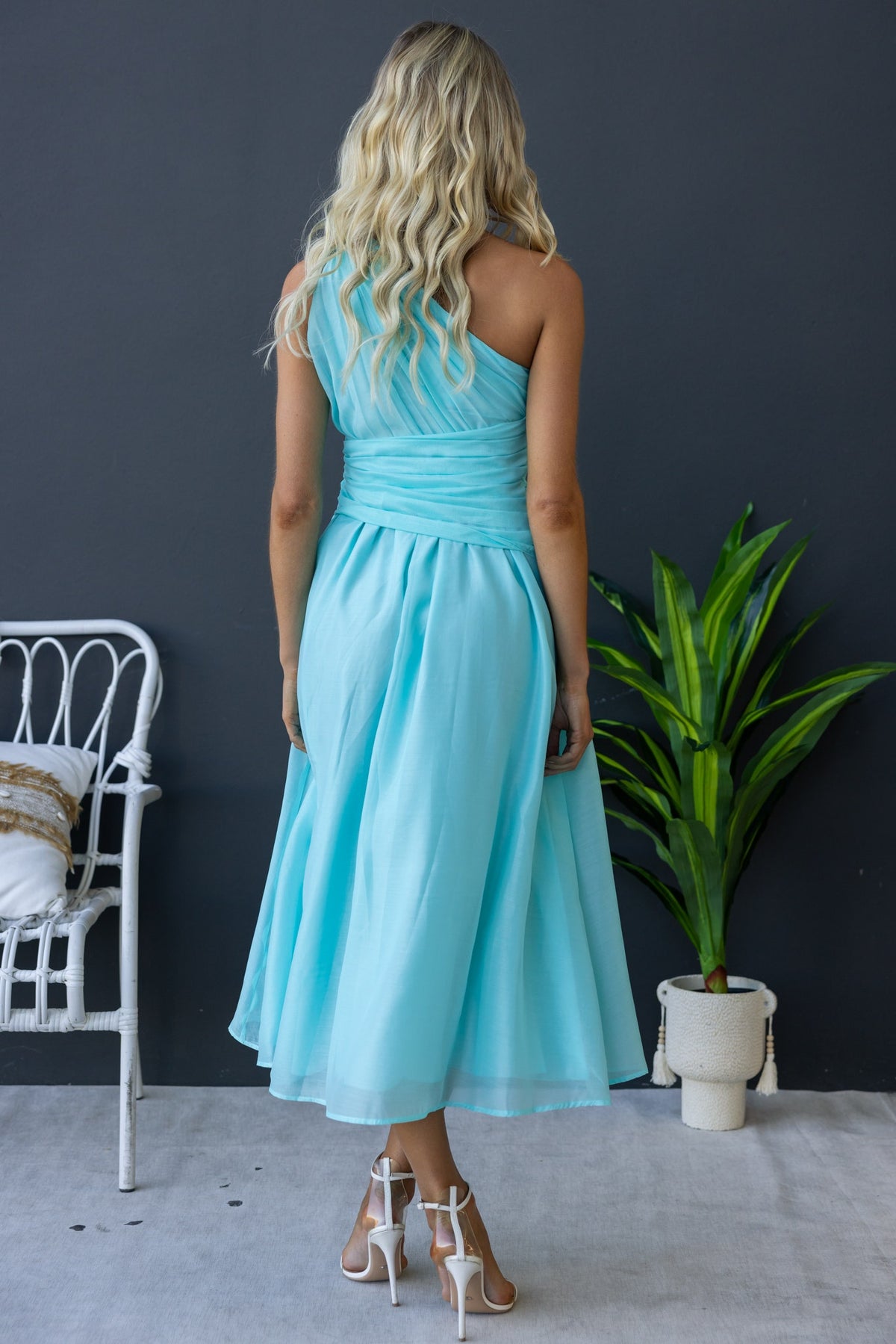 Back view of a woman in an aqua one-shoulder midi dress with knot detail and chiffon fabric.