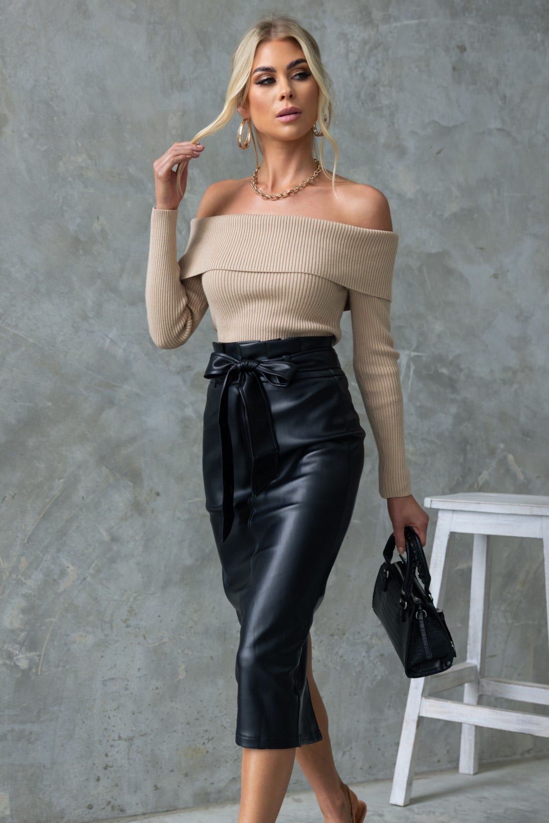 Model wearing Nerida Midi Skirt in black faux leather, off-shoulder beige top, styled with small handbag.