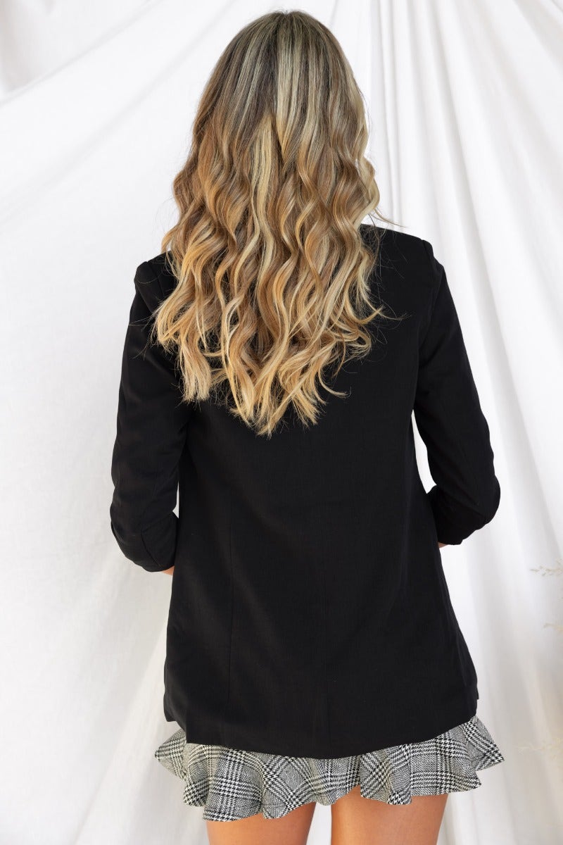 Back view of a woman wearing a black Santino blazer with shoulder pads, styled with a checkered skirt.