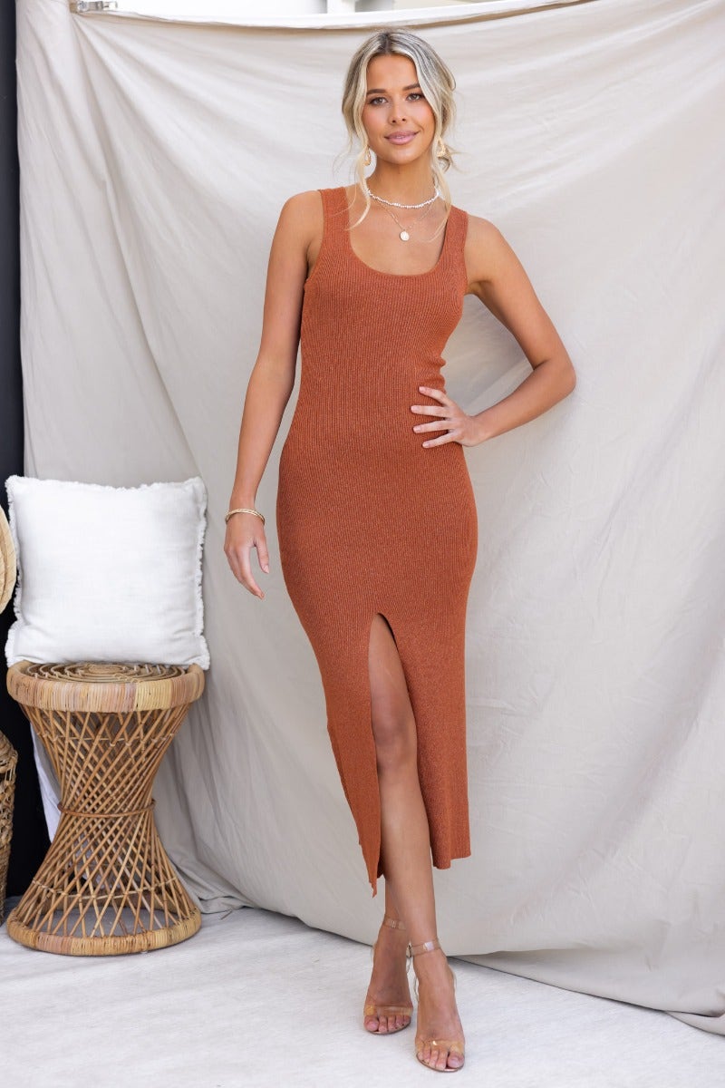 Rata midi dress in rust color, featuring ribbed knit, scoop neckline, and front split detail, styled with strappy heels.