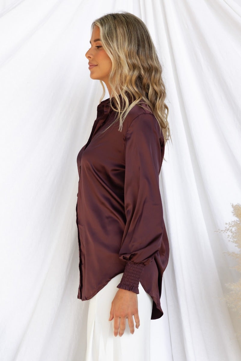Daina Shirt in burgundy satin, featuring a collar and button-up front, showcased on a model from the side.