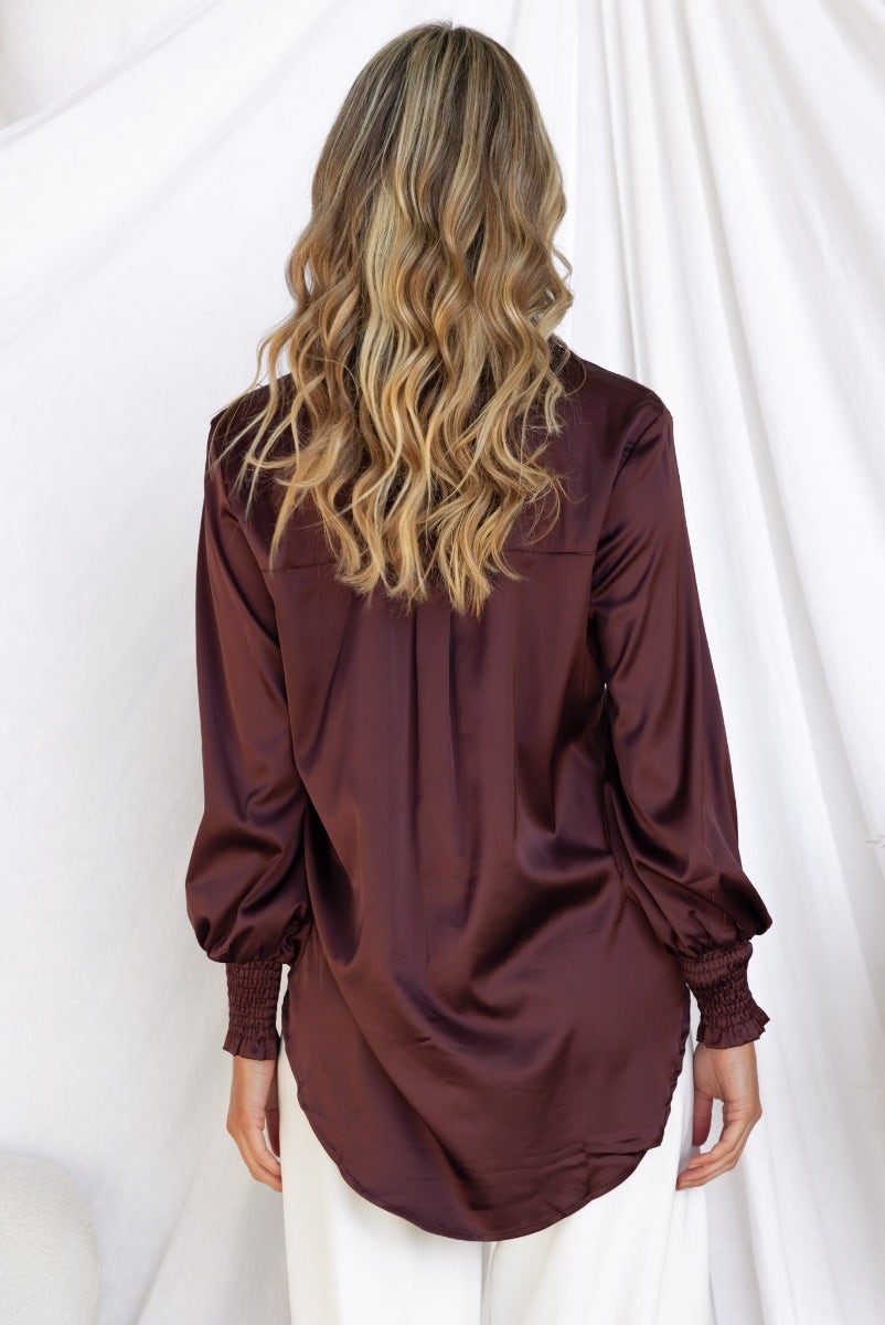 Back view of a woman wearing a burgundy satin Daina shirt with elastic cuffs and relaxed fit.