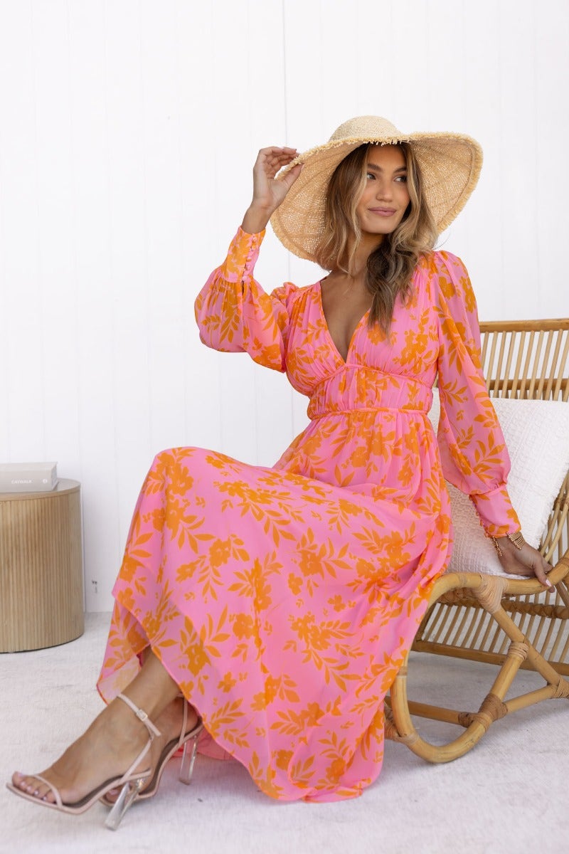 Woman in pink floral Jeanie Maxi Dress with V-neckline and wide-brim hat, sitting comfortably on a chair.