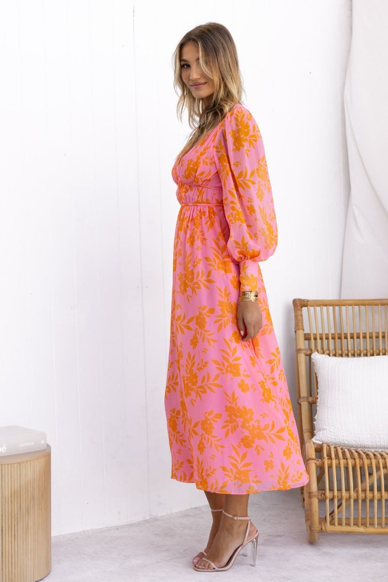 Model wearing a vibrant floral print Jeanie Maxi Dress in pink and orange, showcasing a V-neckline and puffed sleeves.