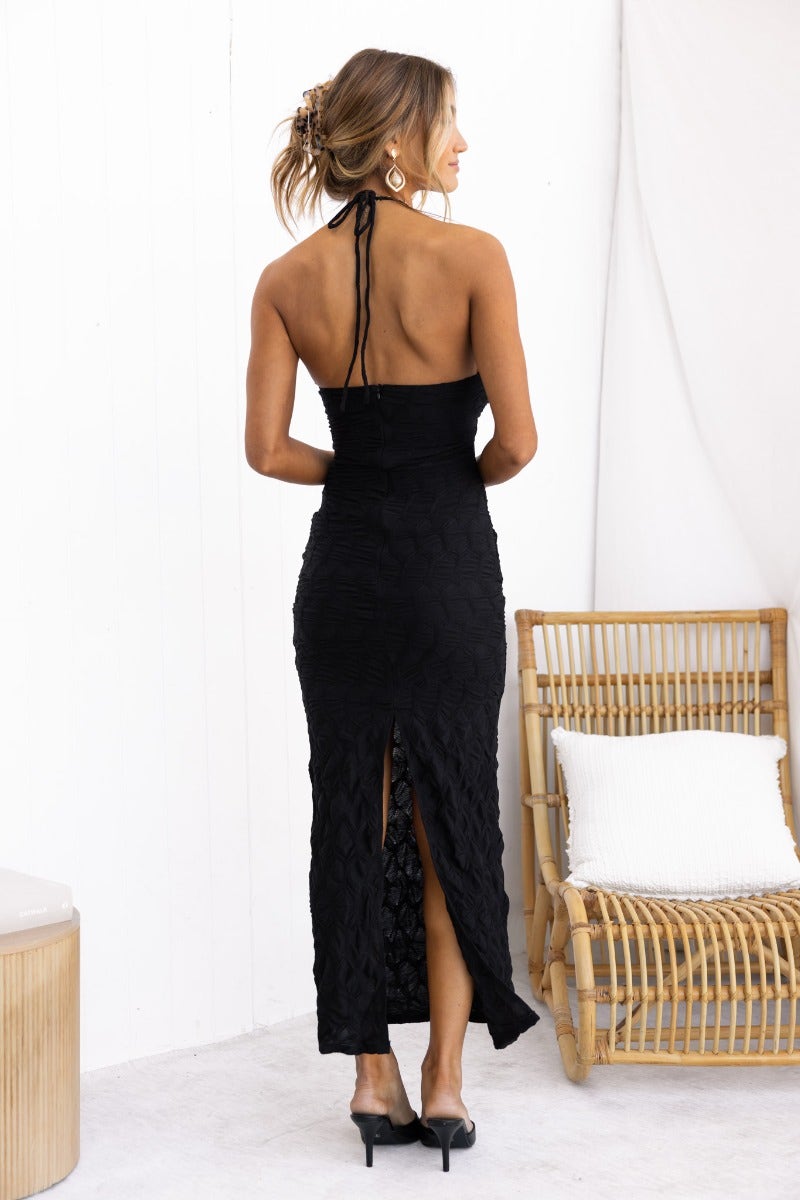 Model wearing the Over Again Maxi Dress, showcasing its black textured fabric and adjustable halter neckline with a back split.