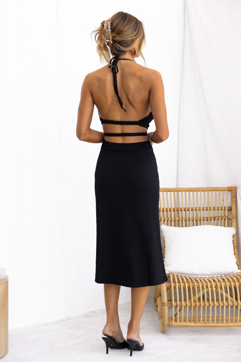 Back view of a woman in a black textured Eyona midi dress with adjustable tie halter neckline and buckle detail.