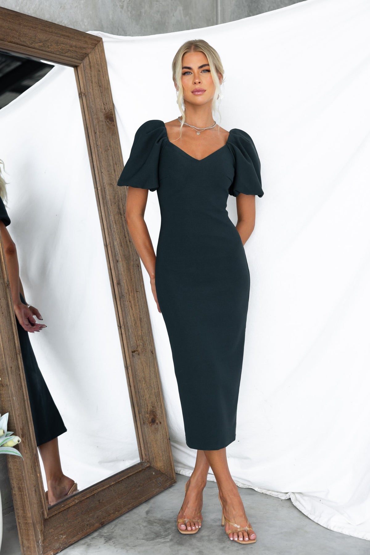 Dark green Raquel Midi Dress with V-neckline, puff sleeves, and back split, styled in a modern setting.