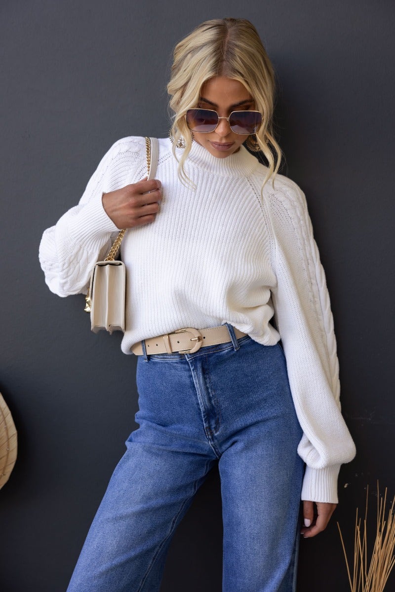 Kallan Jumper in white with high neckline and cable knit sleeves, styled with jeans and a belt.