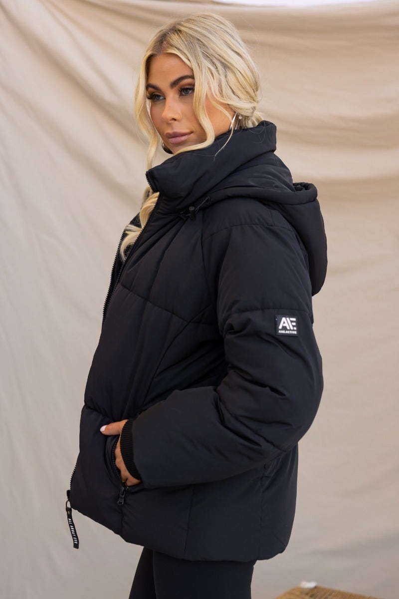 Model wearing a black Delila puffer jacket with a high neckline, removable hood, and zip-up front, styled for a winter look.
