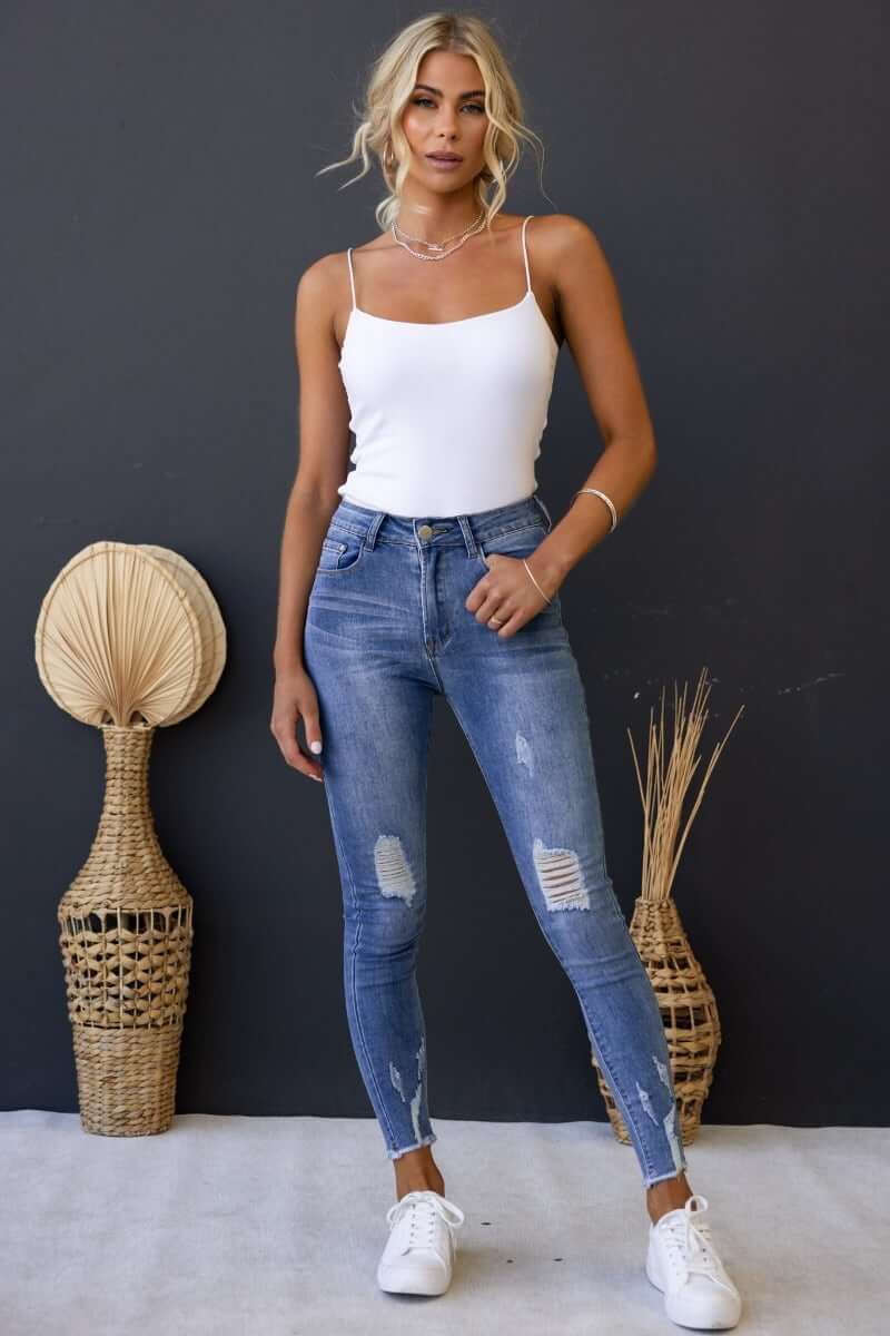 Model wearing Qilli distressed denim jeans with rip details and a raw hem, styled with a white tank top.